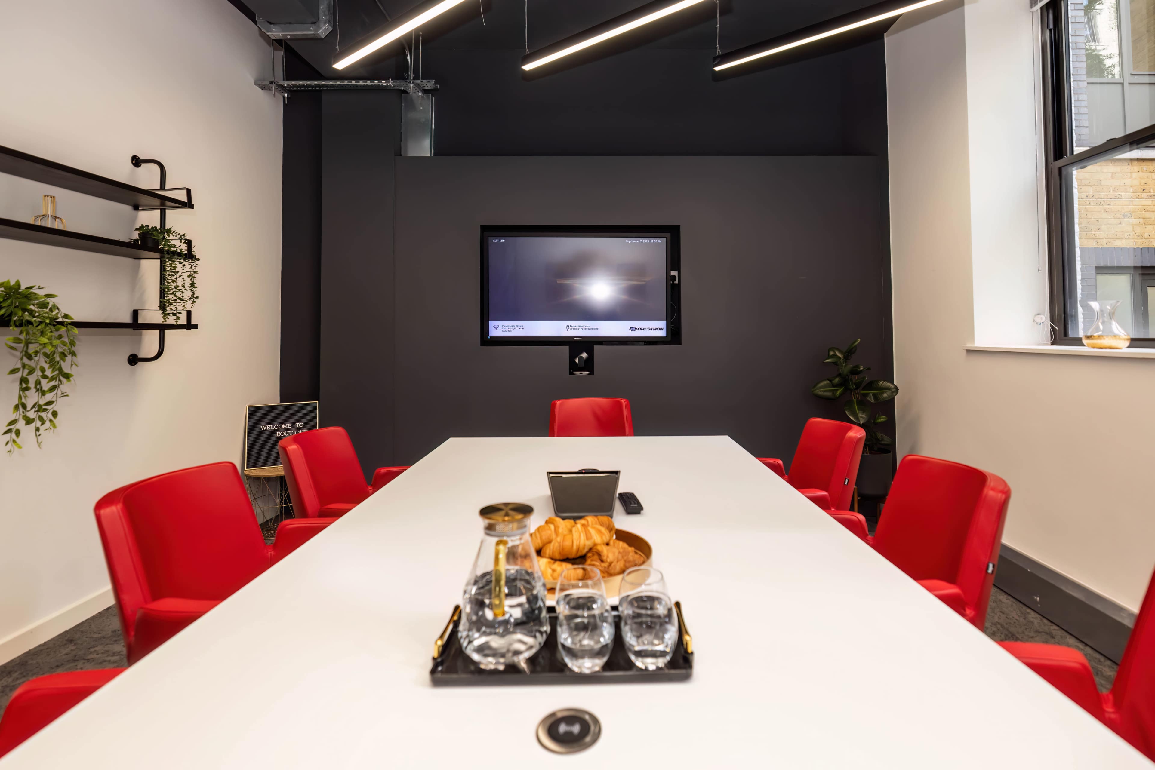 A conference room features a long white table with red chairs, a television mounted on the wall, and refreshments including water and croissants on the table.