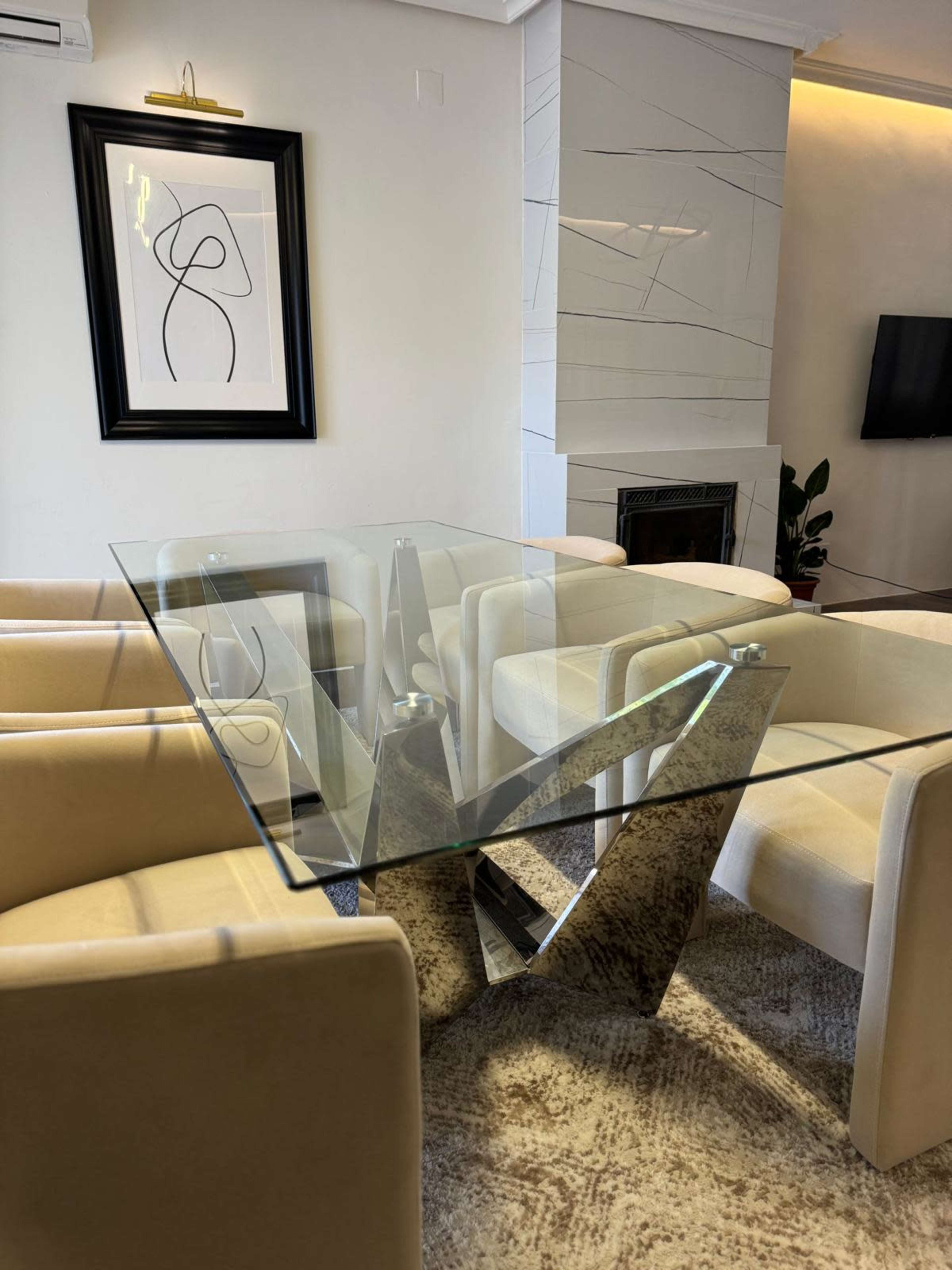 The image depicts a modern living space featuring a glass table with a unique base and beige upholstered chairs arranged around it, alongside a decorative wall with abstract art.