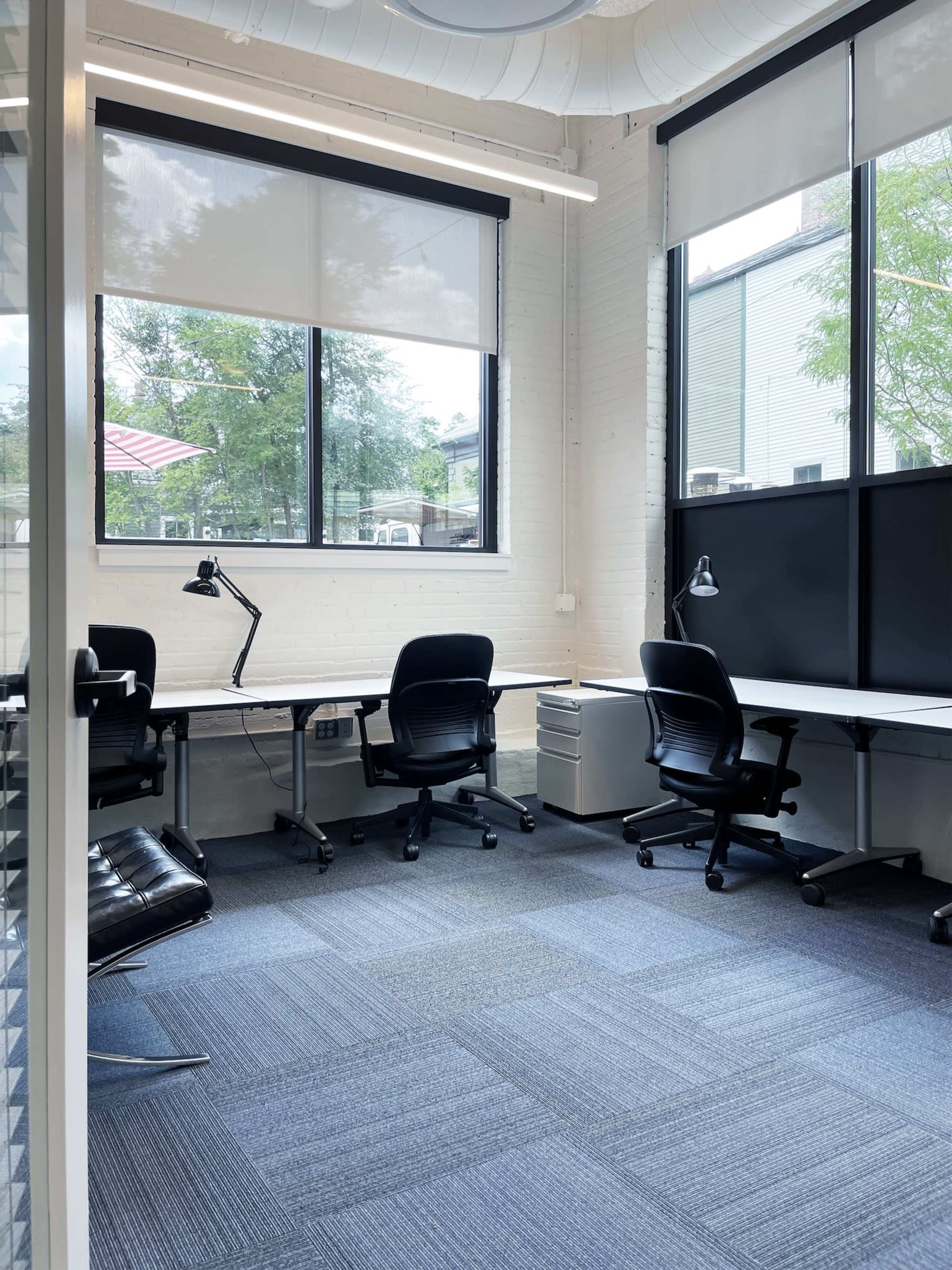A modern office space featuring two desks, office chairs, and large windows with natural light.
