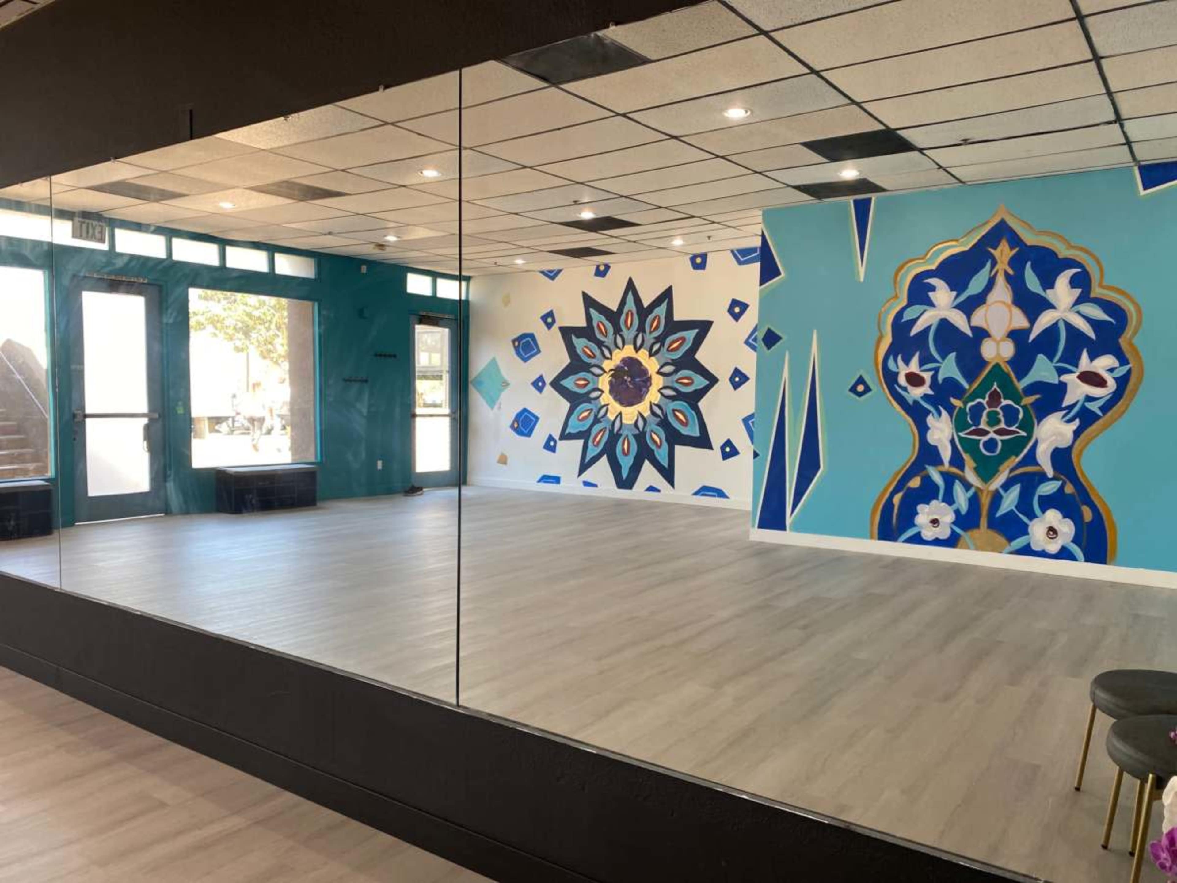 The image shows a spacious room with a mirrored wall and vibrant murals on an opposite wall featuring geometric and floral designs.