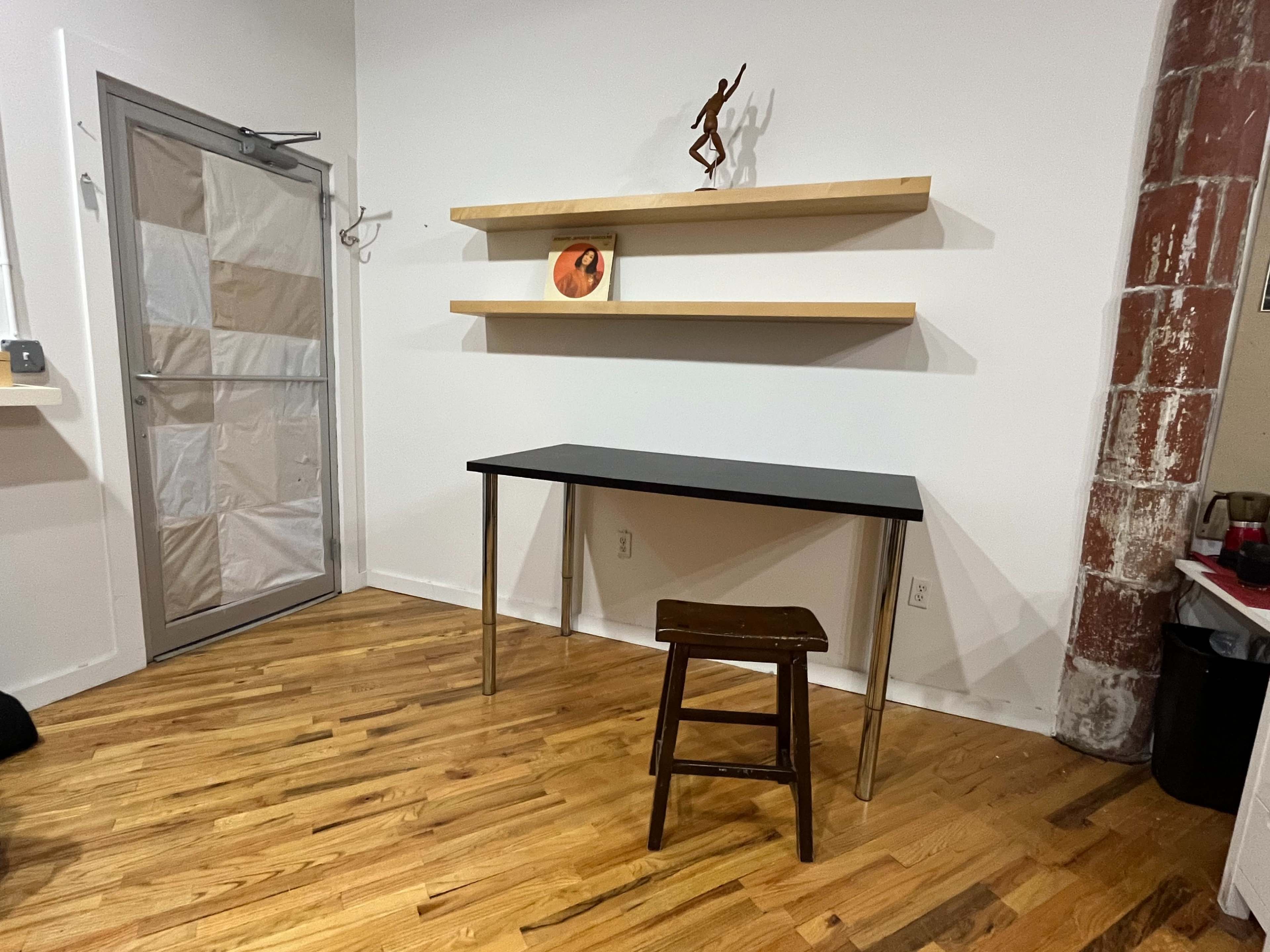 A minimalist room with a wooden floor, featuring a black table, a wooden stool, and a shelf with an object on it.