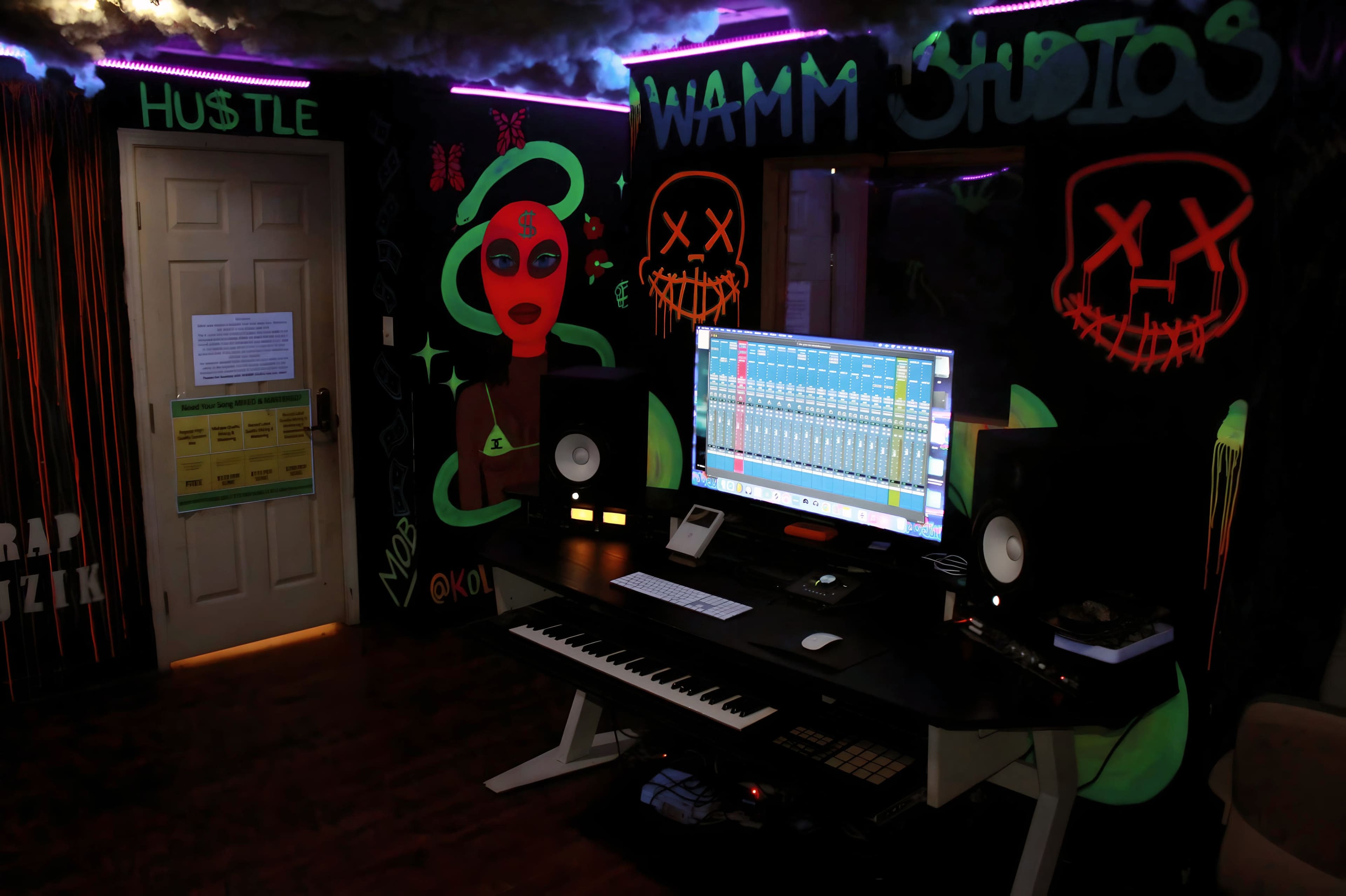 A music production studio features a computer monitor with audio software, a keyboard, and graffiti-style artwork on the walls.