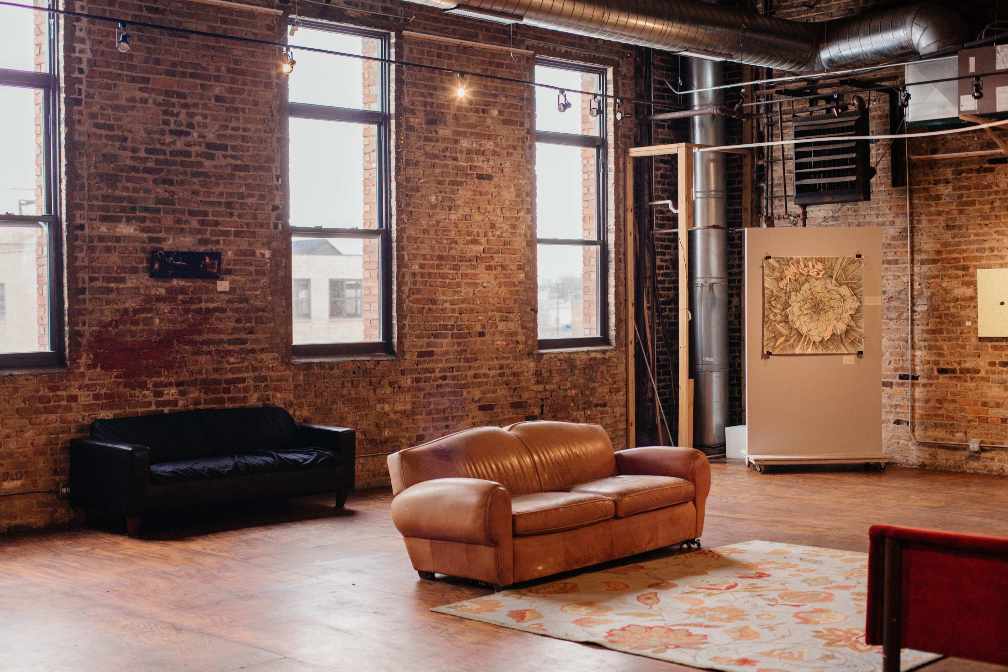 A spacious art gallery with exposed brick walls, large windows, and a mix of furniture, including a brown leather sofa and a black couch on a wooden floor.