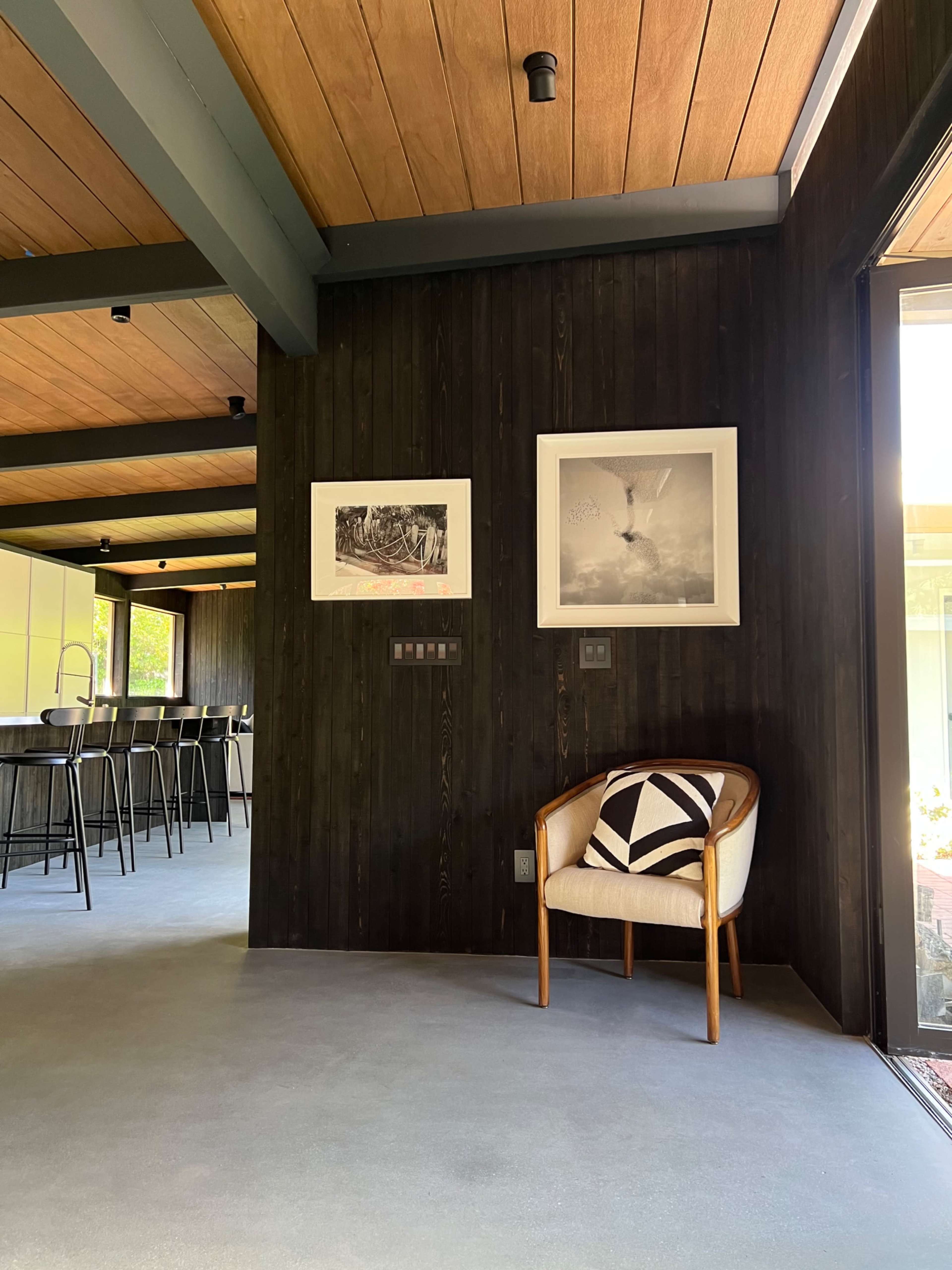 A corner of a modern interior with a wooden chair and framed artwork on the black wall, leading to a bright kitchen space.