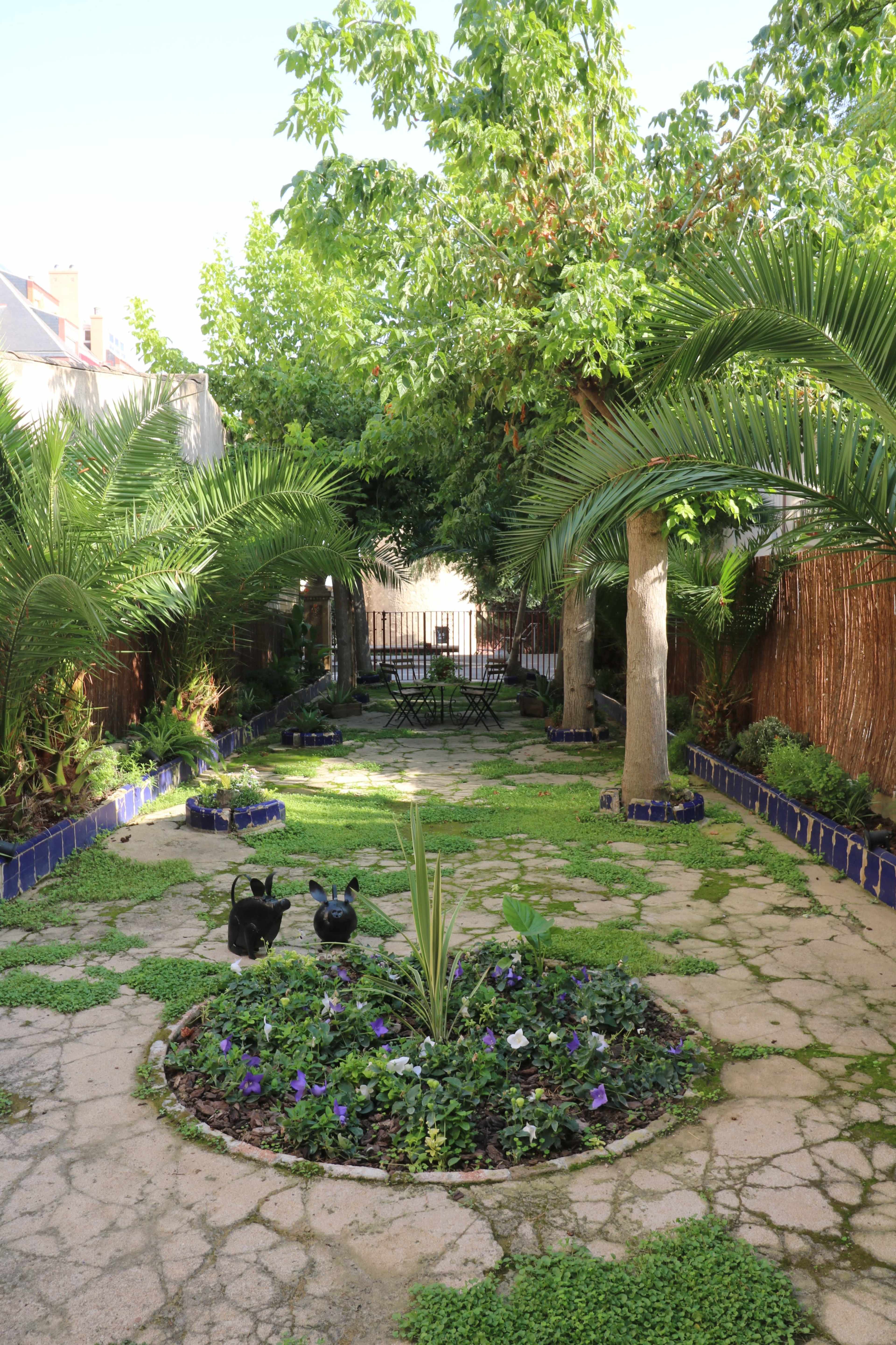 A green garden with palm trees, a circular flowerbed, and a seating area at the back surrounded by lush vegetation.