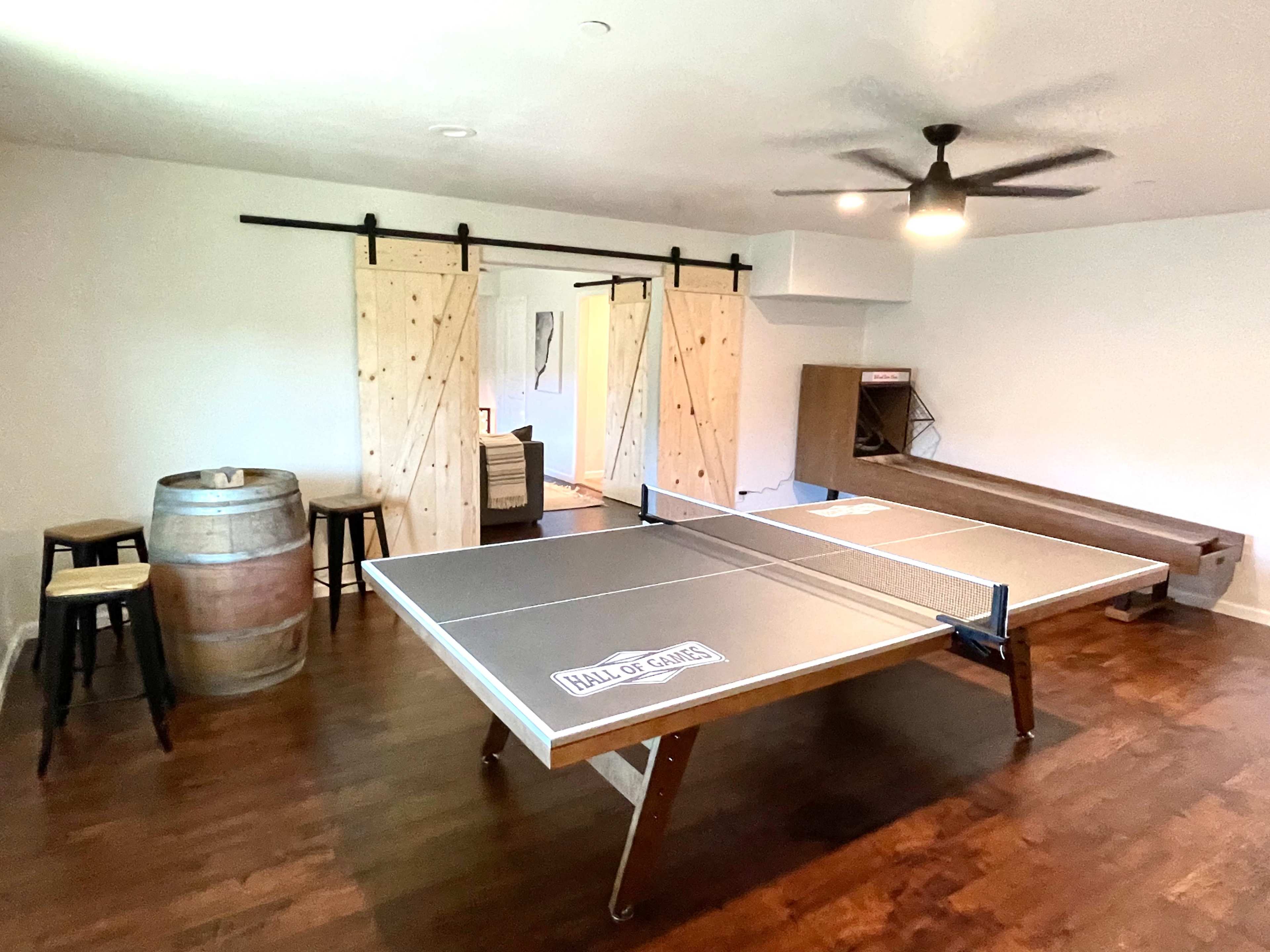 The image shows a recreational room featuring a ping pong table, a wooden barrel, stools, and sliding barn doors leading to another area.