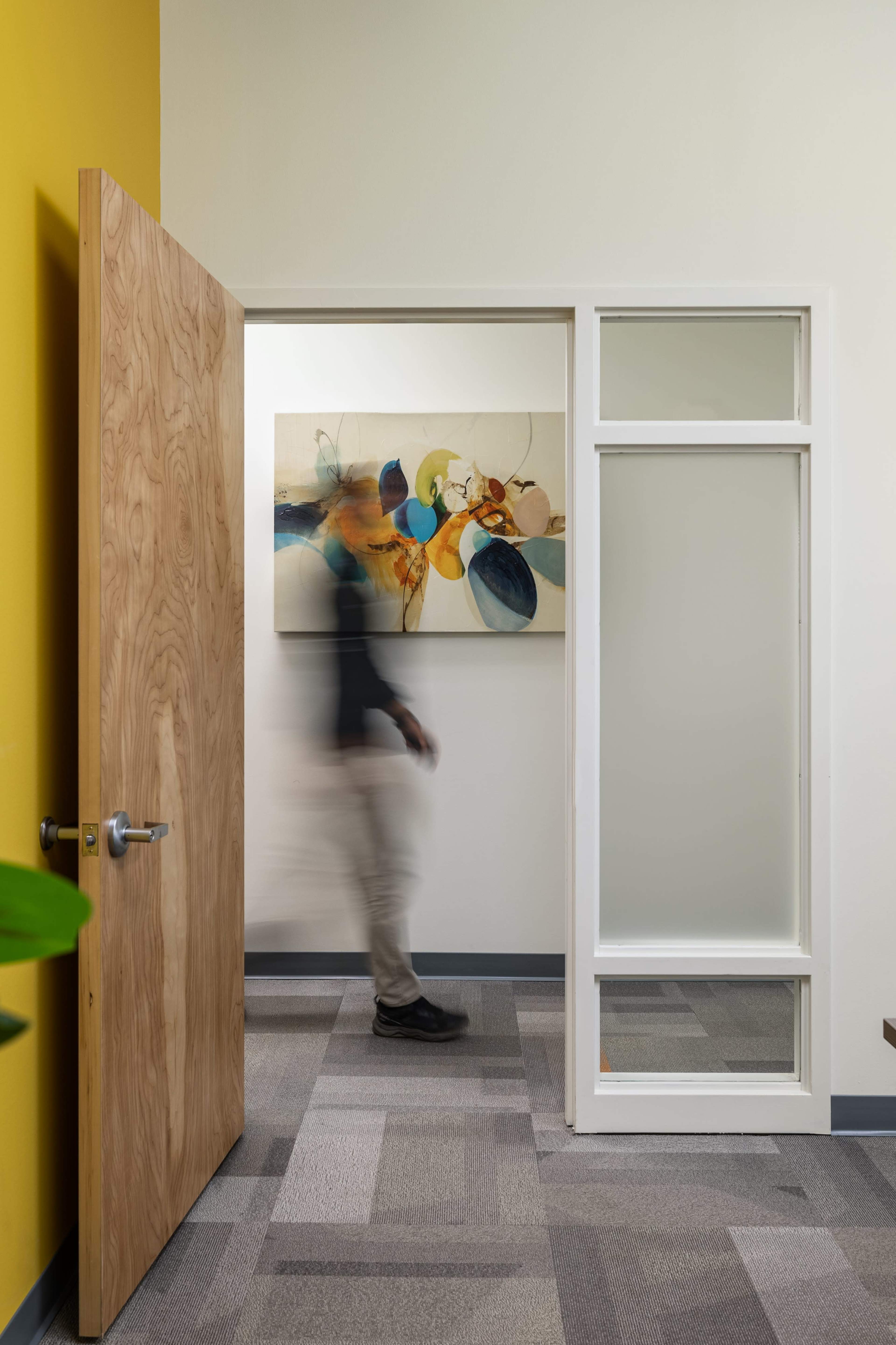 A person walks past an open door with a wooden frame and a colorful abstract painting on the wall in a hallway.