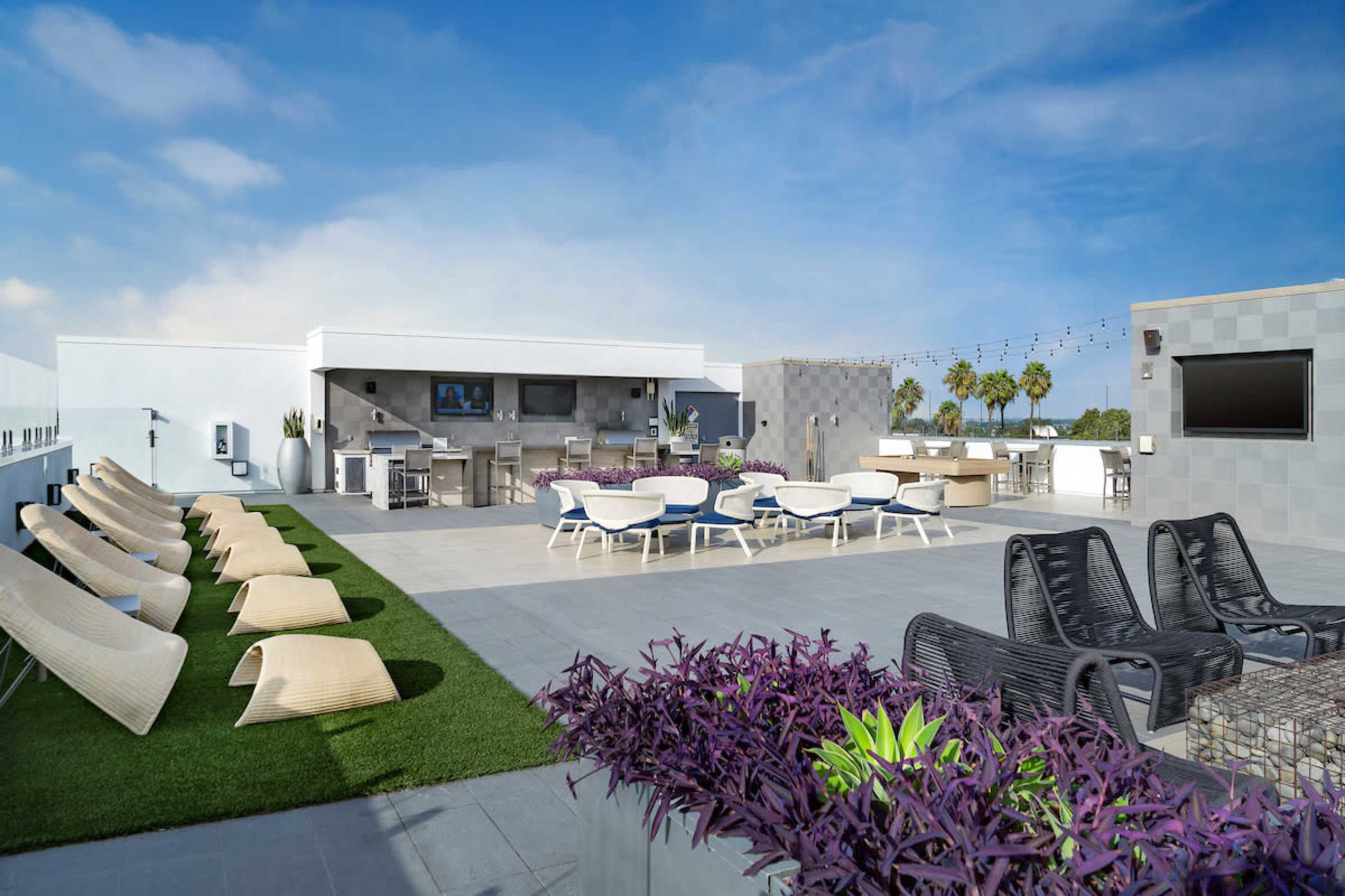 The image shows a modern rooftop terrace featuring lounge chairs, dining tables, an outdoor kitchen area, and landscaping with purple plants.