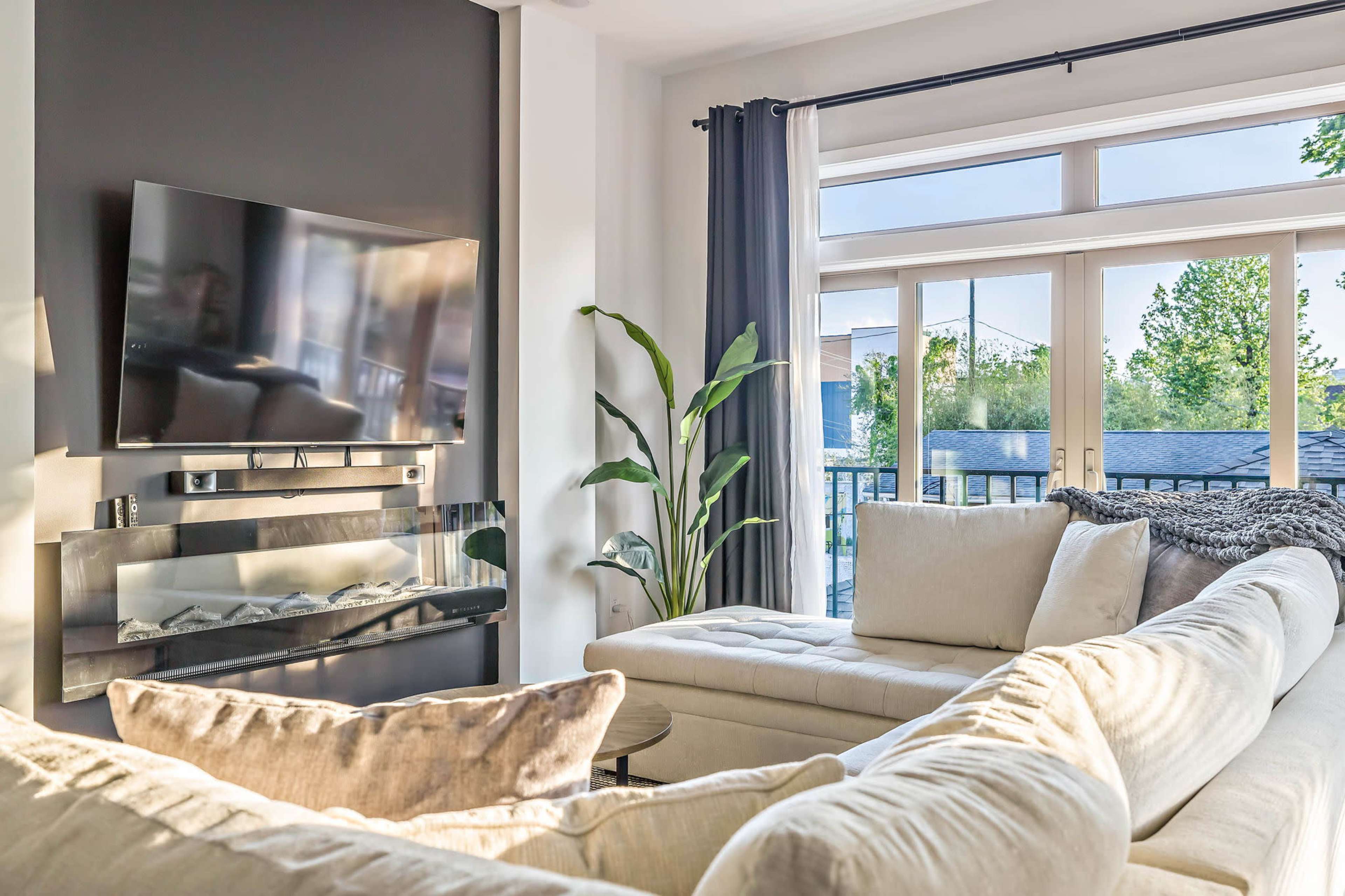 A modern living room features a large sectional sofa, a mounted television, and a wall of windows with natural light illuminating the space.