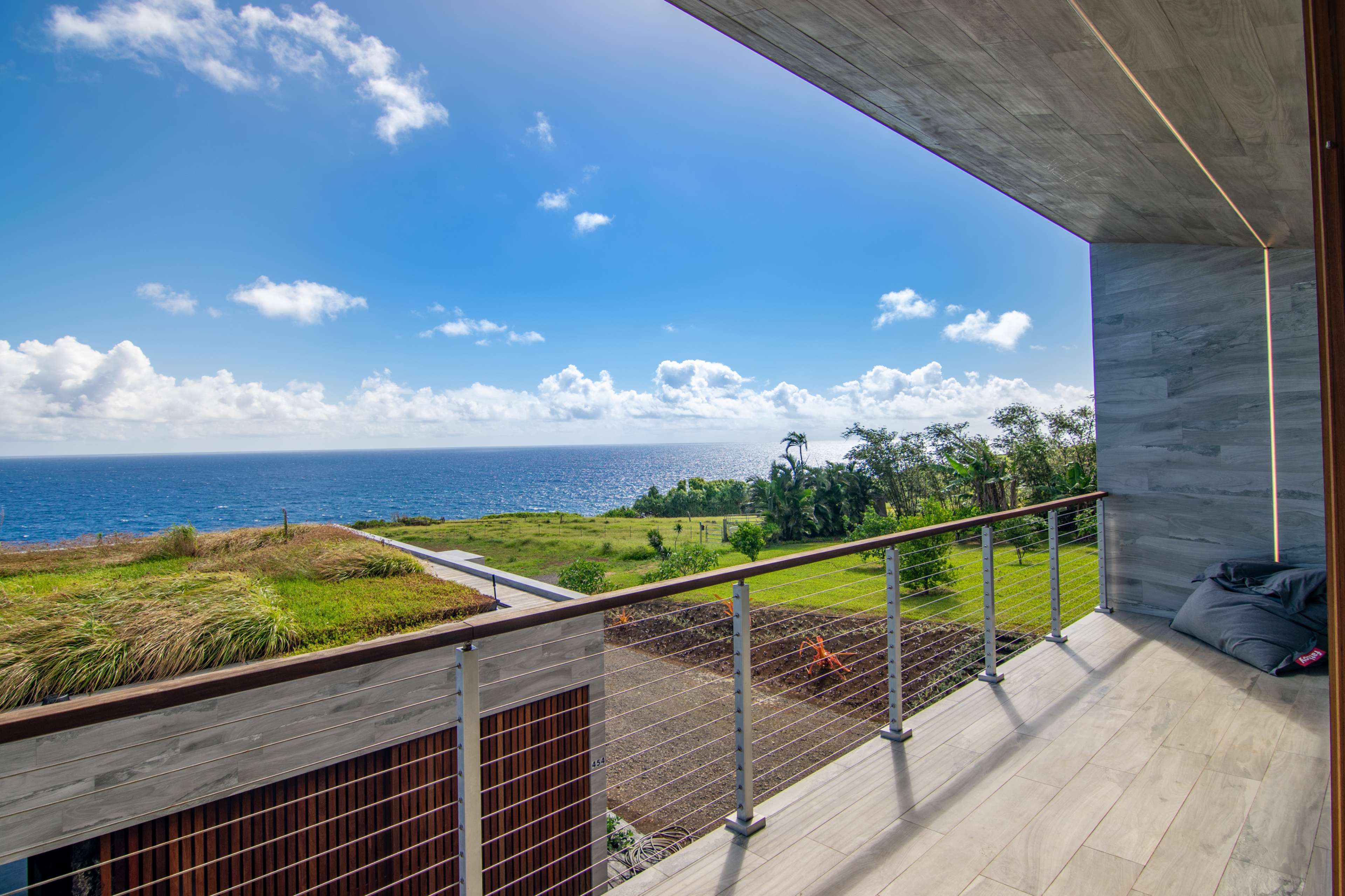 A balcony with a railing overlooks a vast ocean and lush greenery under a blue sky with scattered clouds.