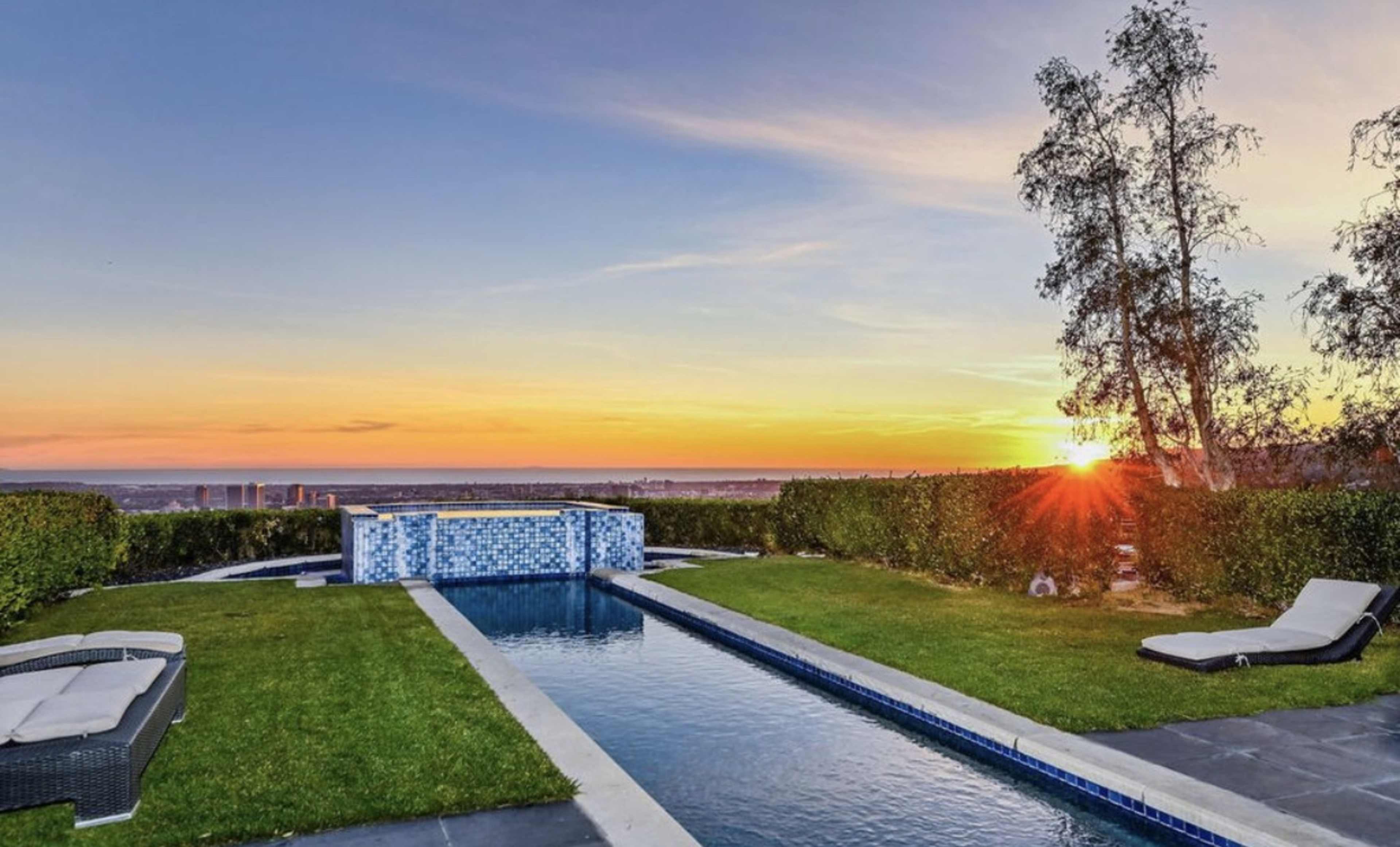 A swimming pool with a sunset backdrop, surrounded by lush greenery and a stone patio.