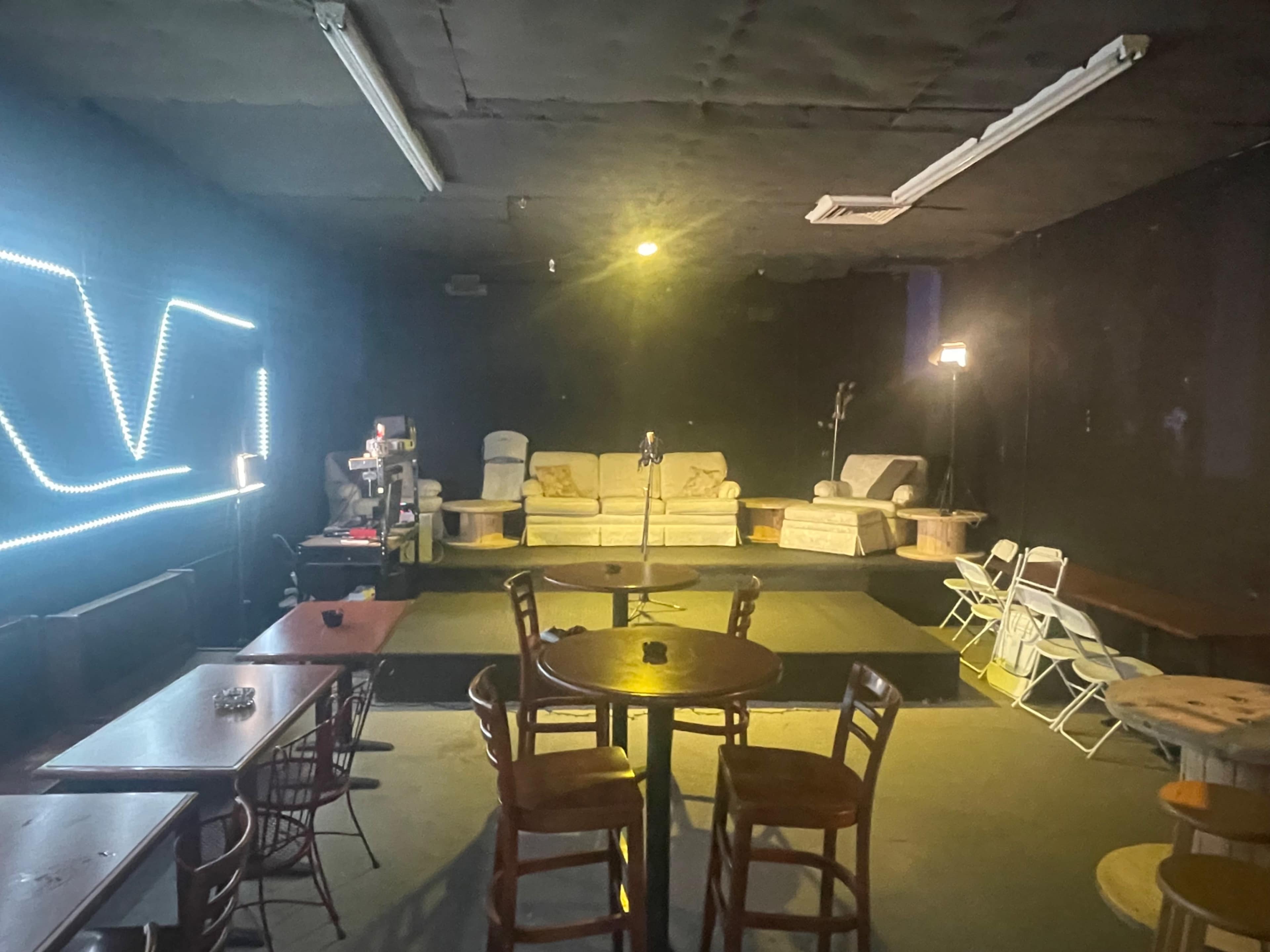 The image shows a dimly lit indoor space with a small stage, a microphone at the center, seating areas including sofas and tables, and a neon light fixture on the wall.