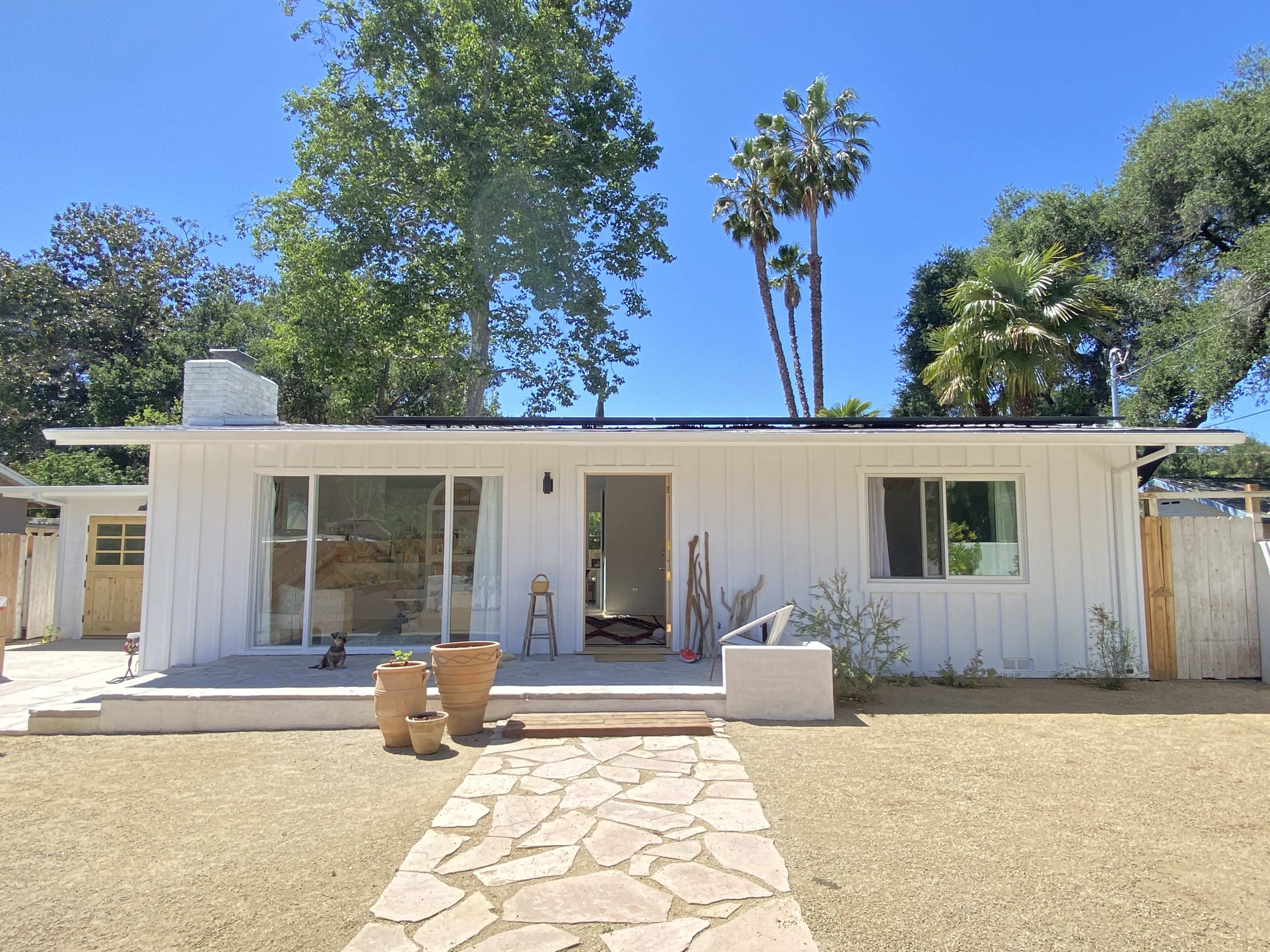 A single-story white house with a stone pathway leads to the entrance, surrounded by trees and palm plants.