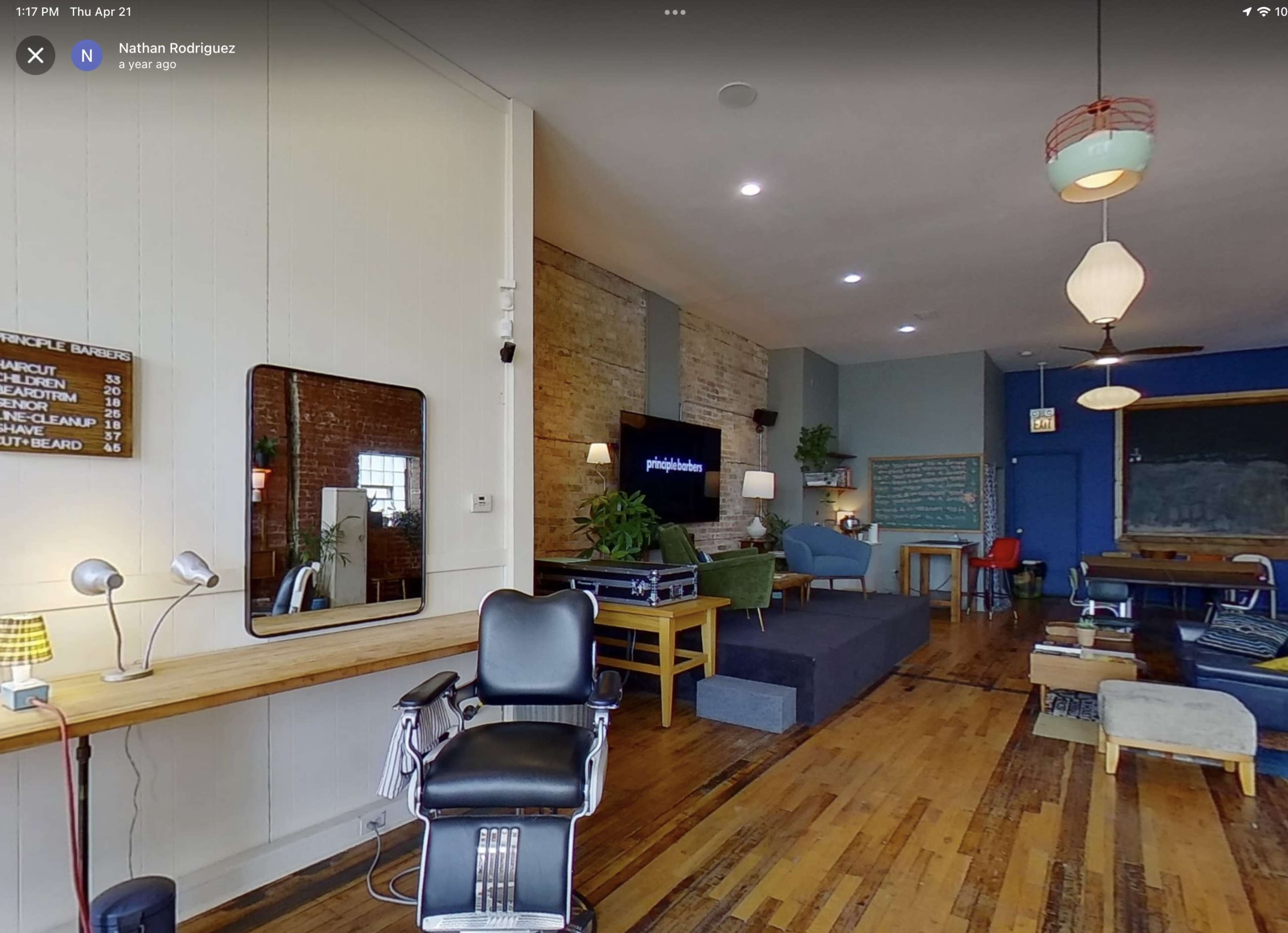 The image shows a modern barbershop interior with a classic barber chair, wooden flooring, a large mirror, and a mix of contemporary and vintage decor elements.