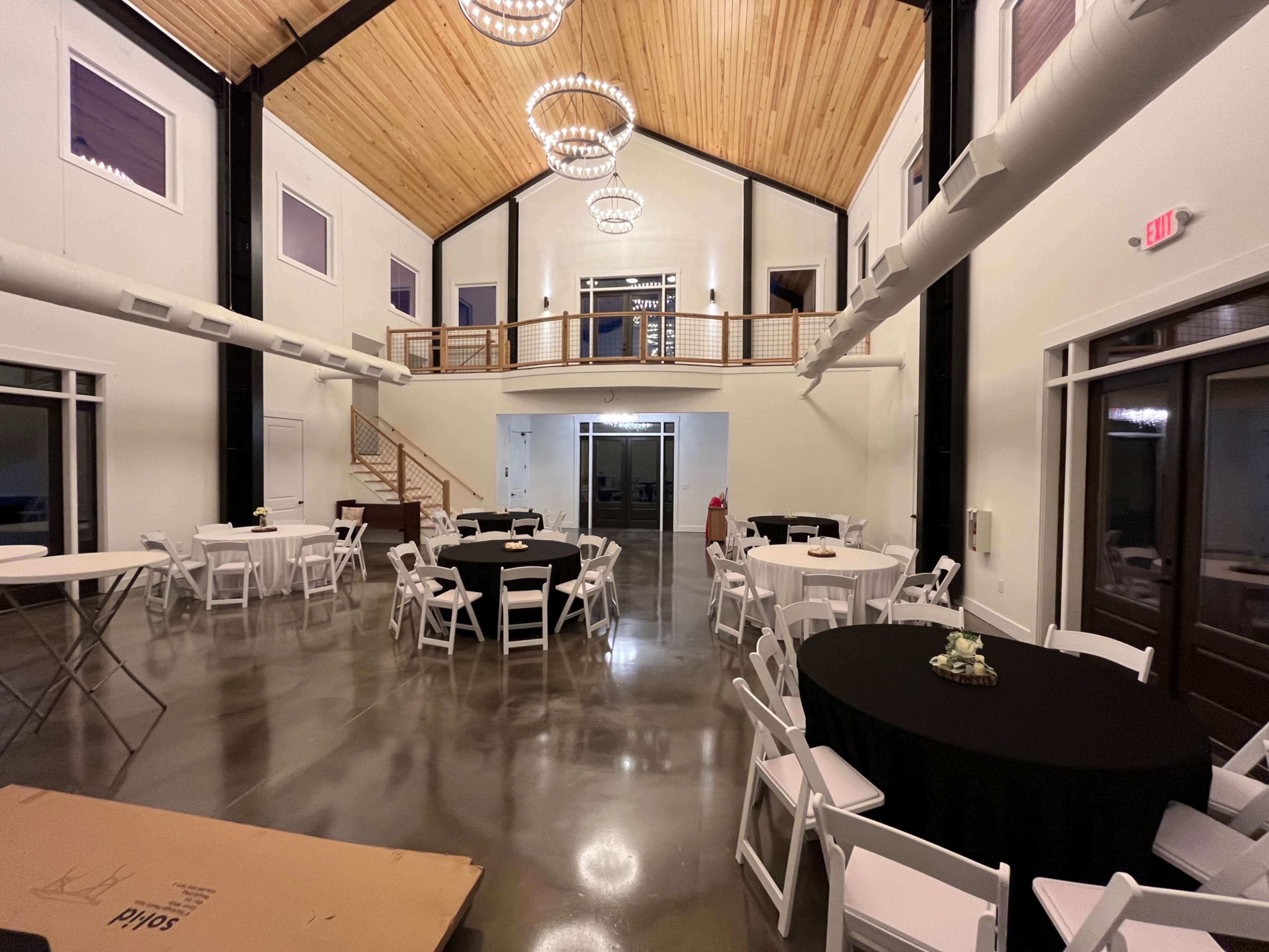 A spacious indoor venue featuring round tables with black tablecloths, white chairs, and large windows, with wooden beams and a staircase leading to an upper level.