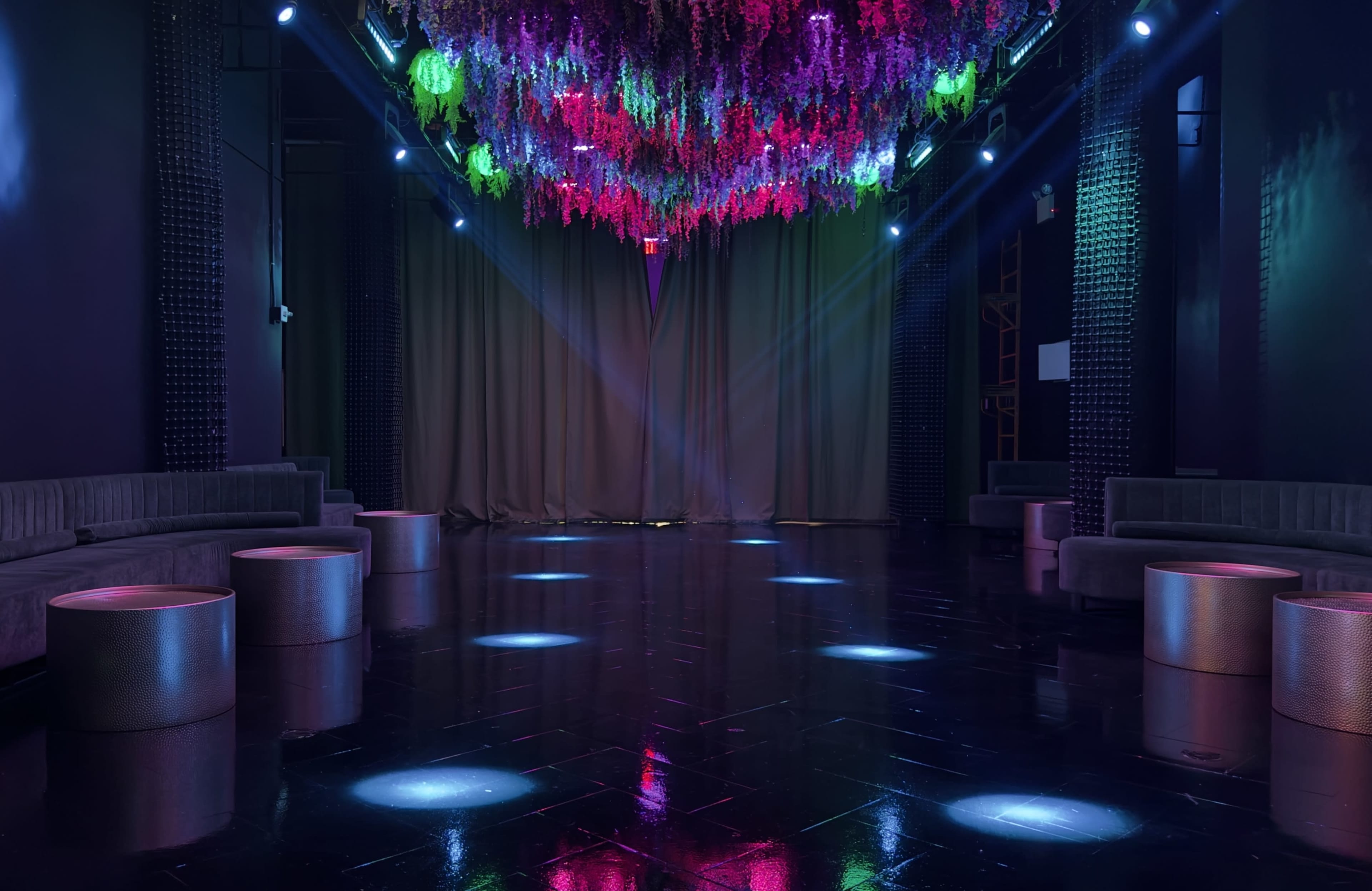 The image shows a dimly lit venue with a glossy black floor, illuminated by colorful lights and adorned with hanging floral decorations.