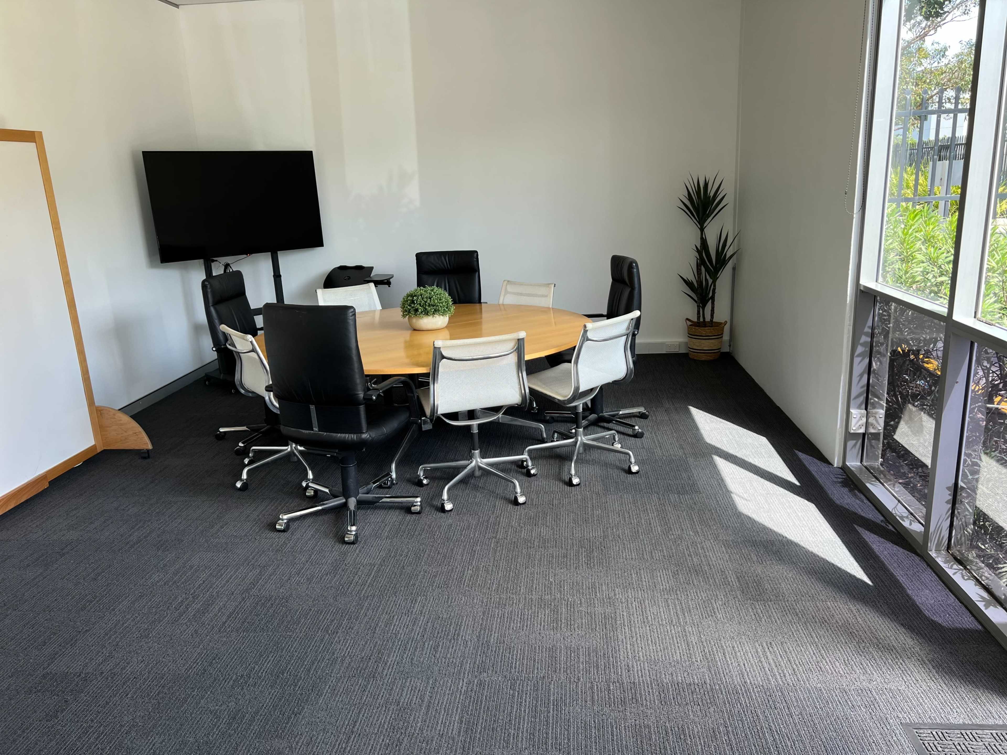 A conference room features a round wooden table surrounded by eight chairs, with a television mounted on the wall and a plant in the corner.
