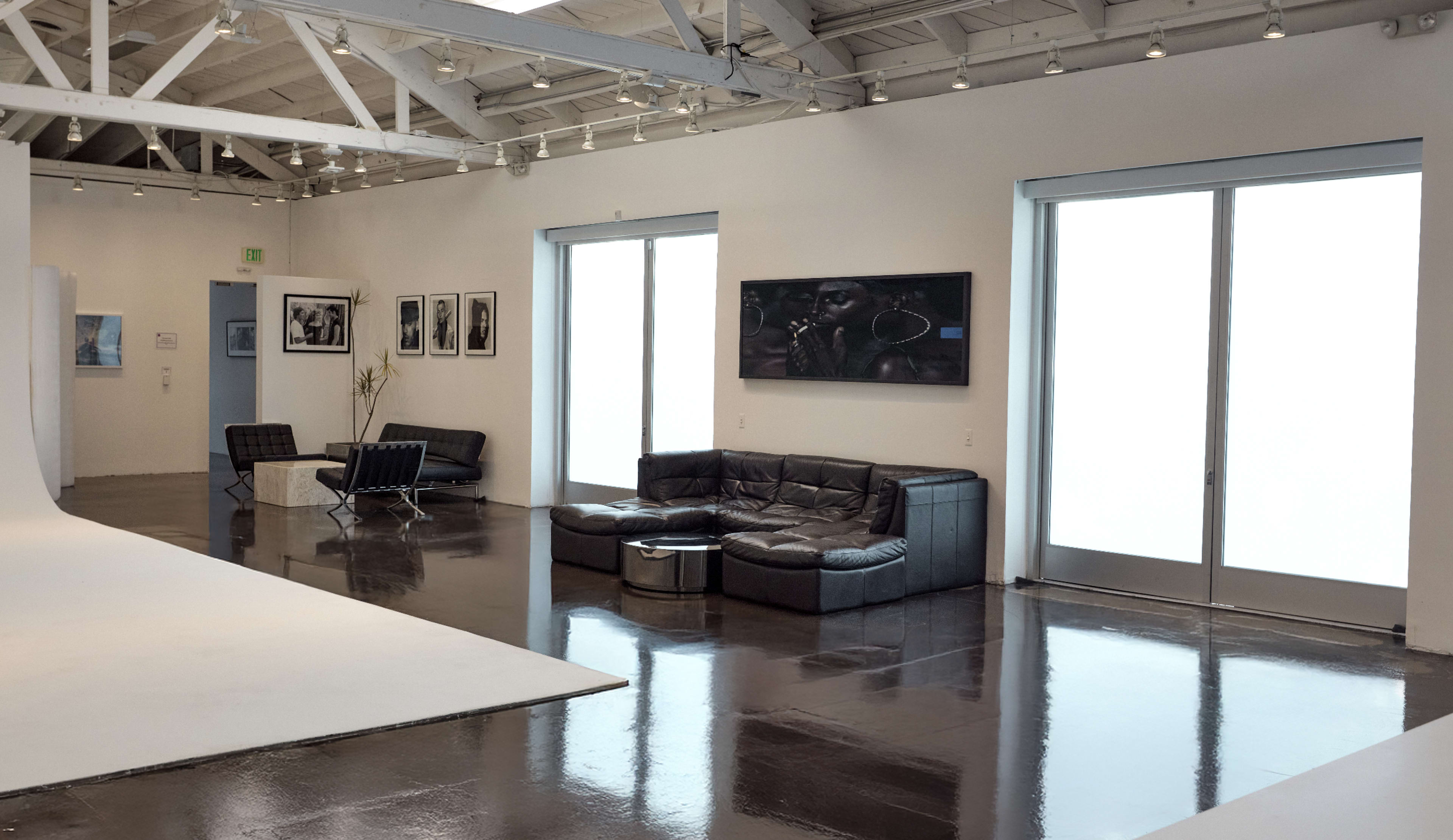 The image shows a modern art gallery with a sleek interior featuring a black leather sofa, a glass table, and framed photographs on the walls.