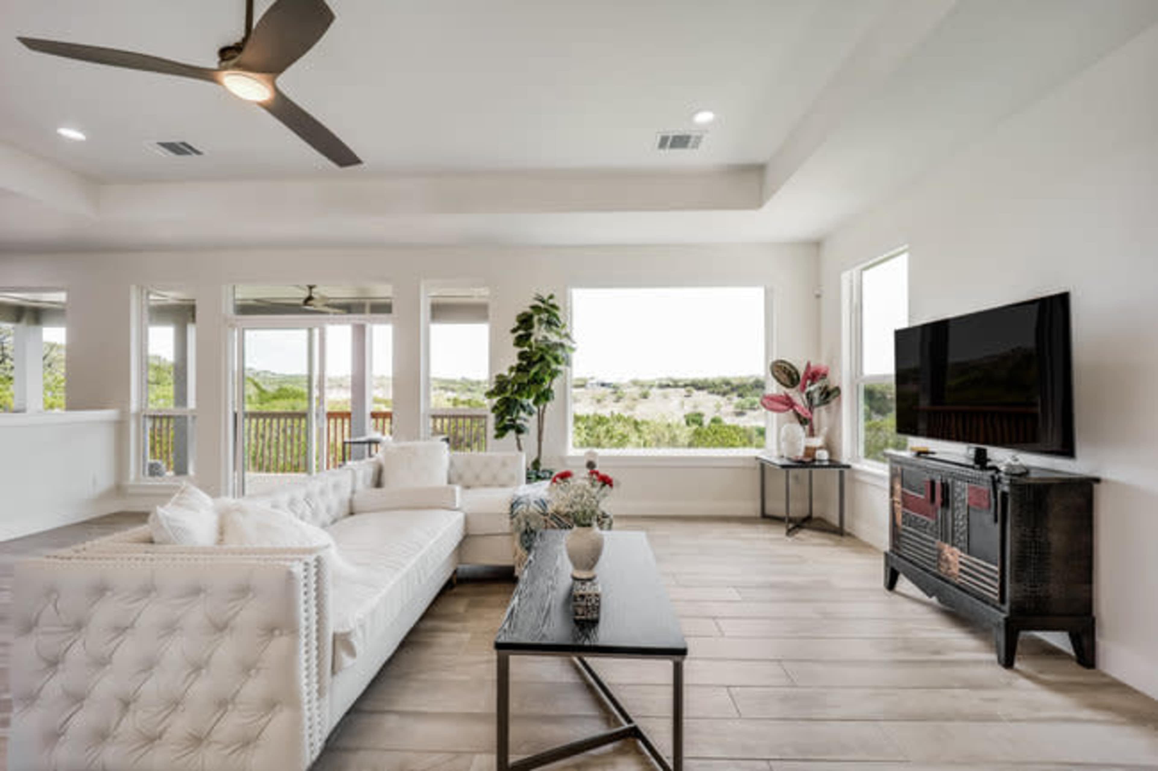 The image shows a modern living room featuring a white tufted sofa, a coffee table, a television, and large windows that offer a view of the outdoor landscape.