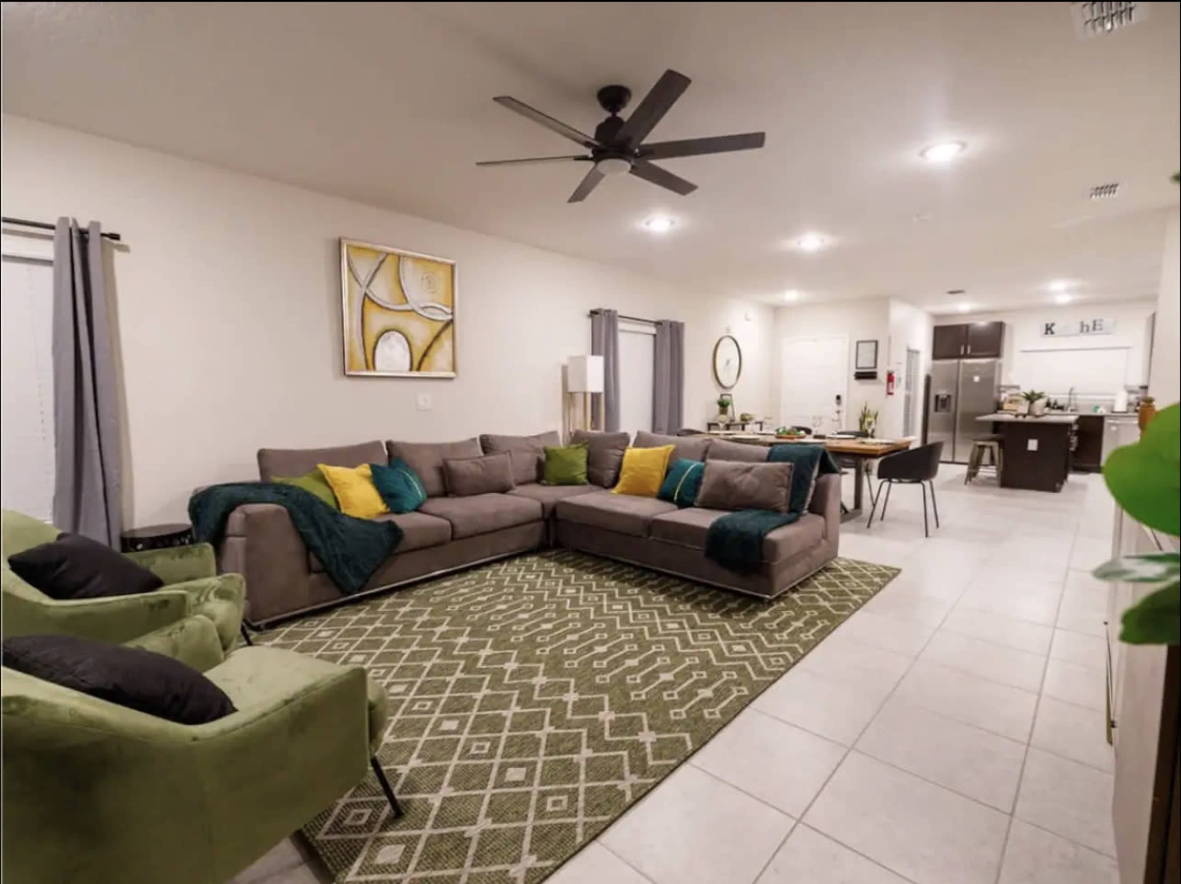 A spacious living area features a large gray sectional sofa with green and yellow cushions, a patterned rug, and a connected dining space with a kitchen in the background.