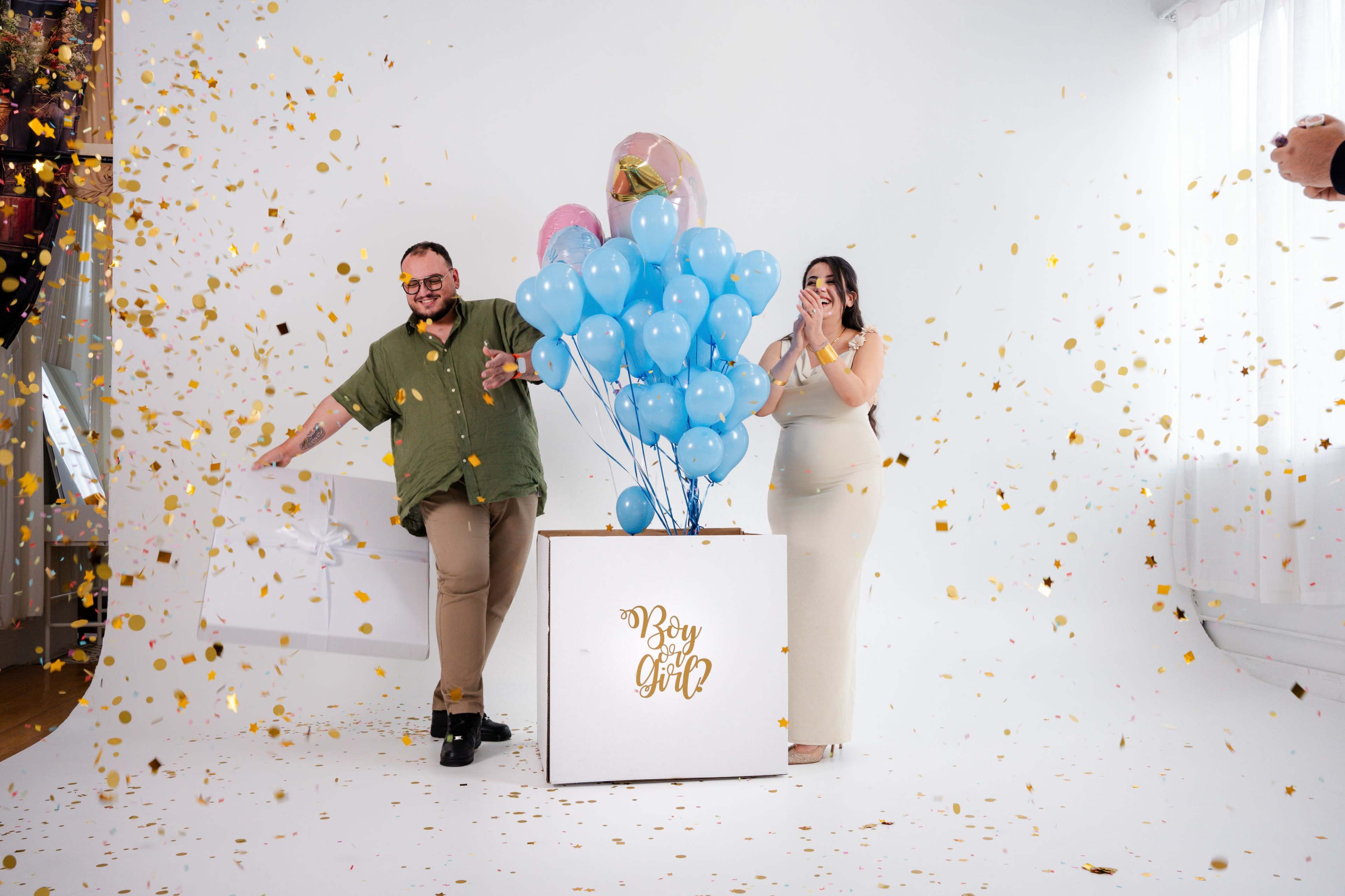 A man and a woman celebrate in a studio setting as blue balloons and confetti are released from a large gift box labeled "Baby Girl."