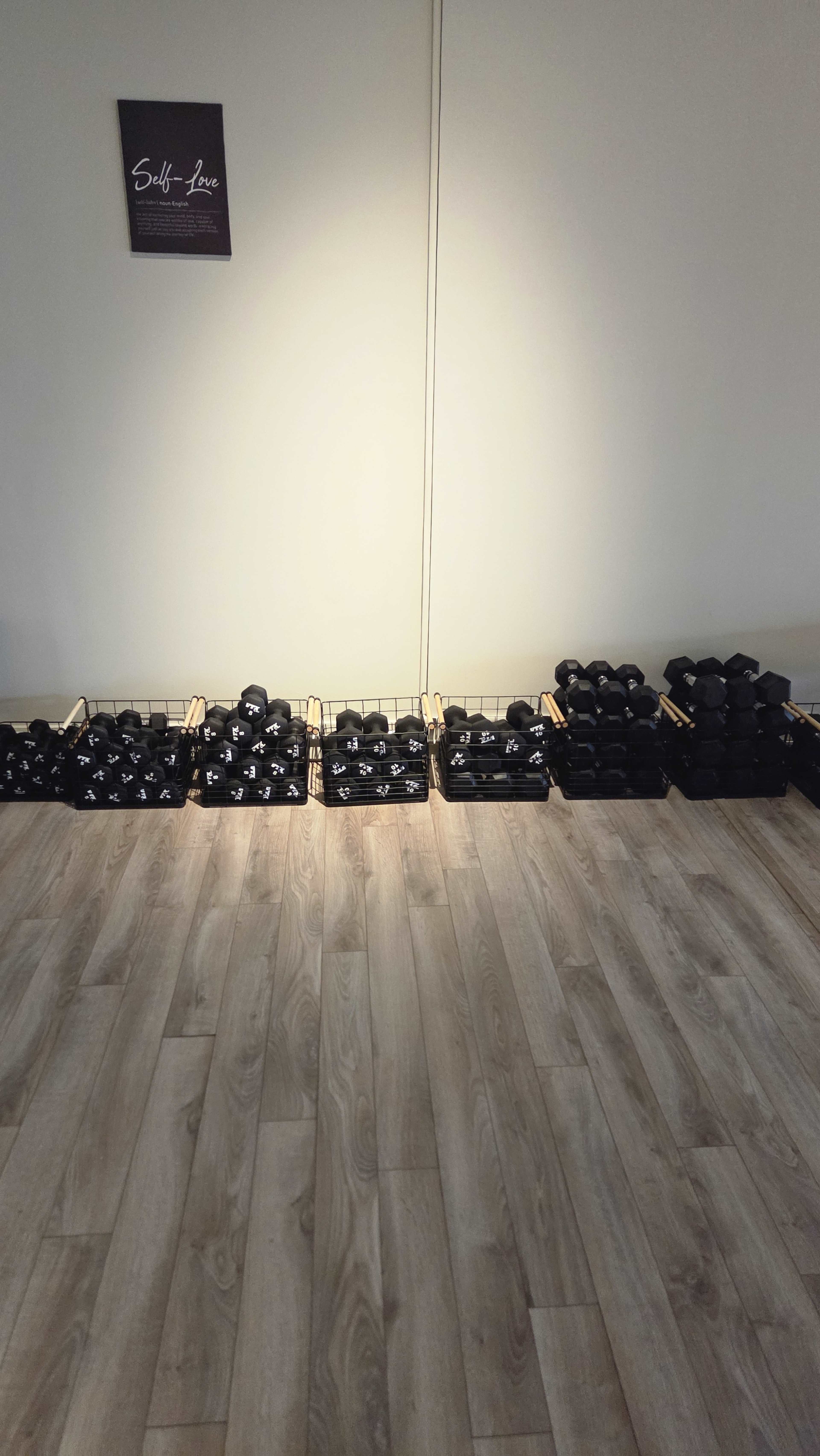 The image shows a neatly organized row of dumbbells on a wooden floor, with a wall in the background.