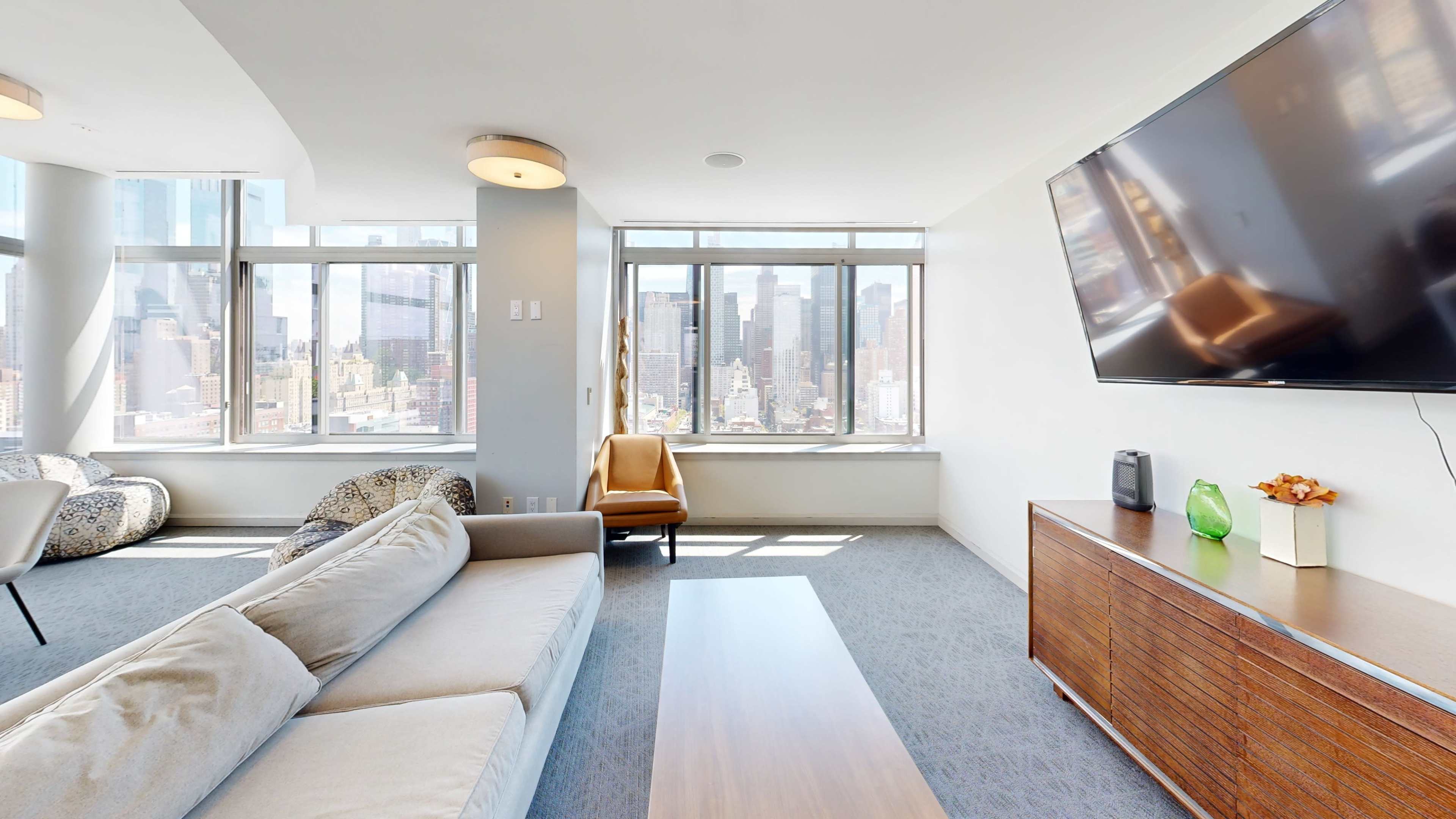The image shows a modern living room with large windows revealing a city skyline, featuring a sofa, a wooden coffee table, a TV mounted on the wall, and a side chair near the window.