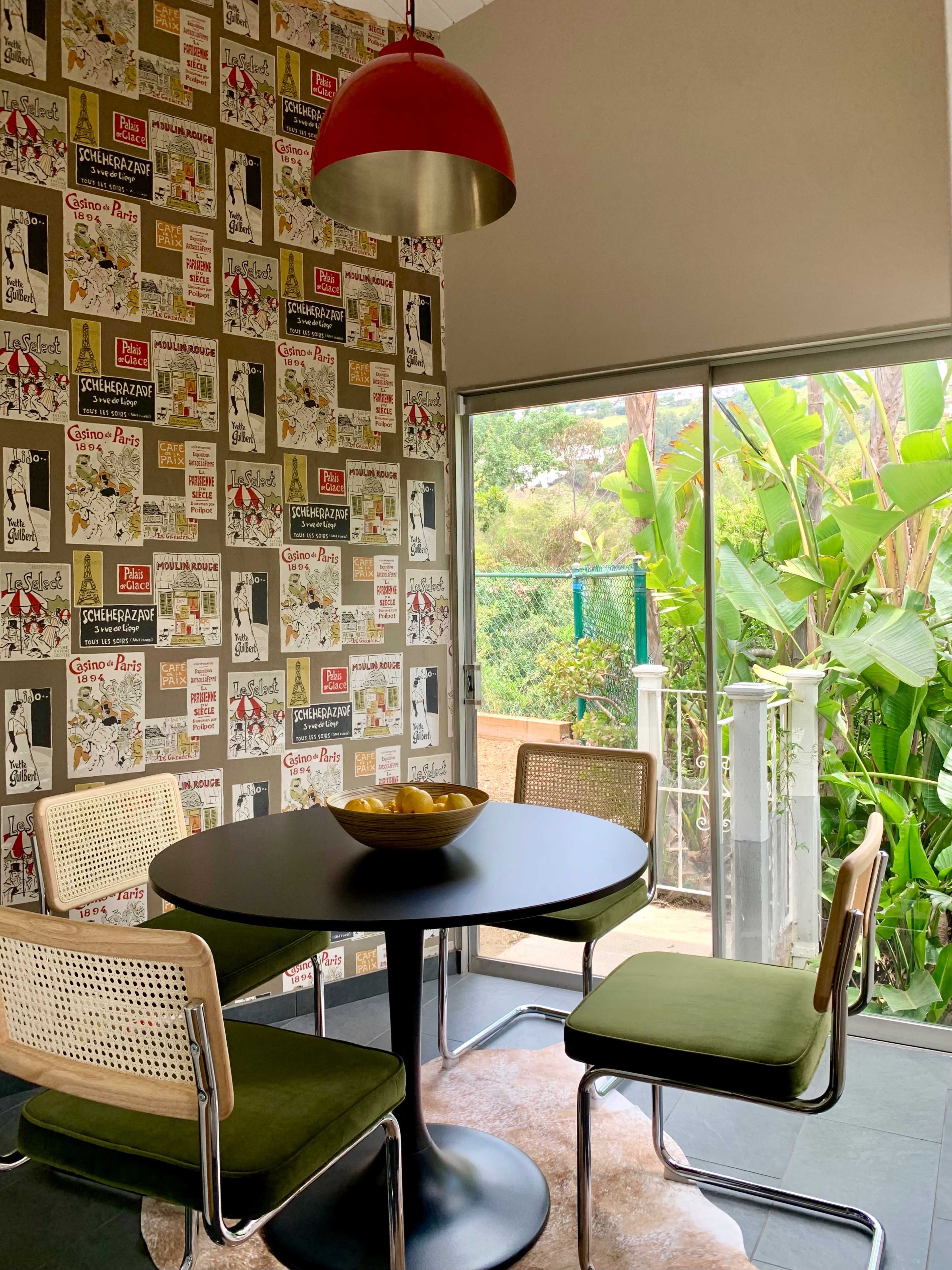 A modern dining area features a round black table with four chairs and a bowl of fruit, surrounded by wallpaper and a large window overlooking greenery.