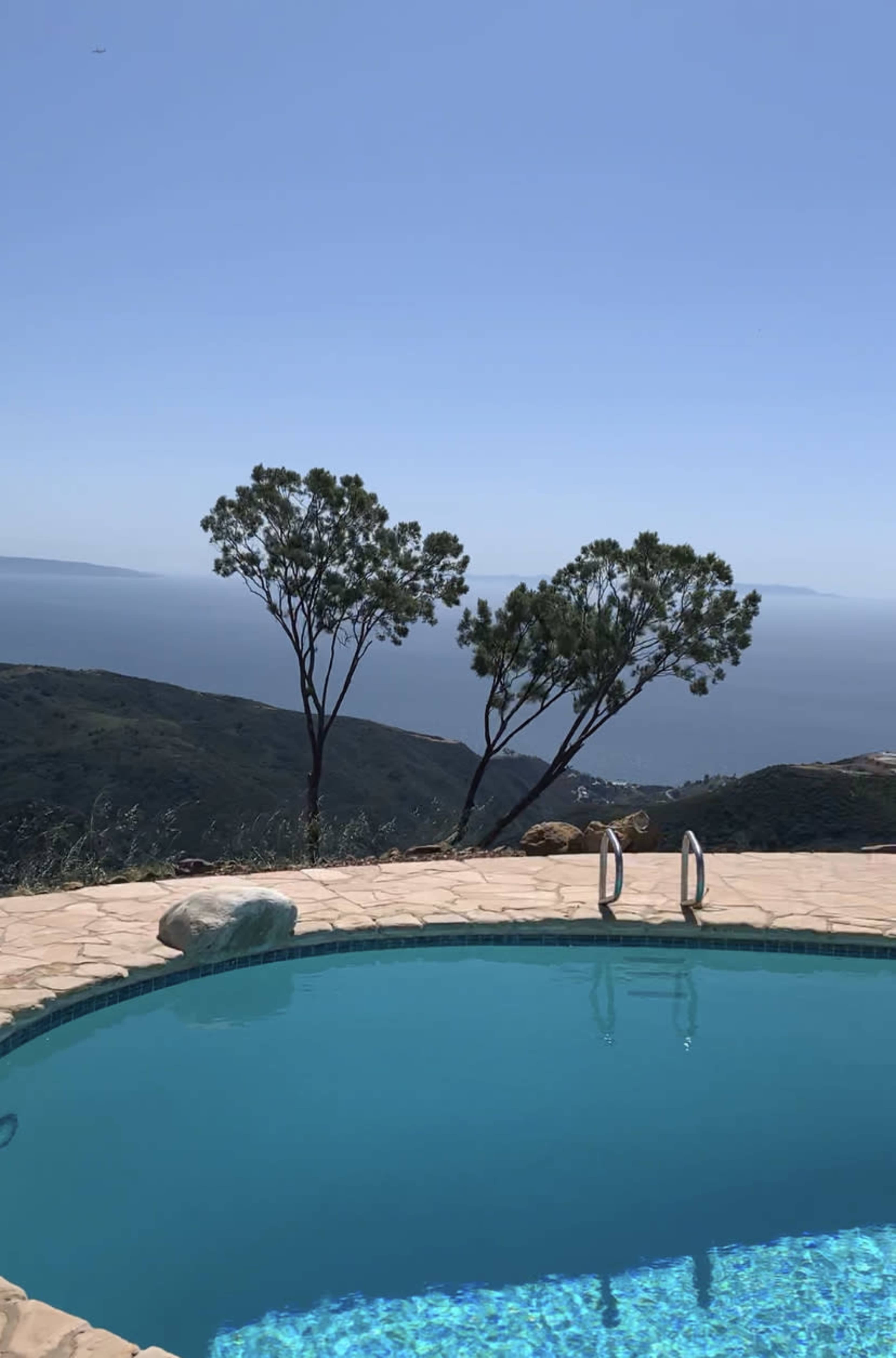A clear blue swimming pool is situated on a stone patio overlooking a distant ocean and rolling green hills, with two trees nearby.