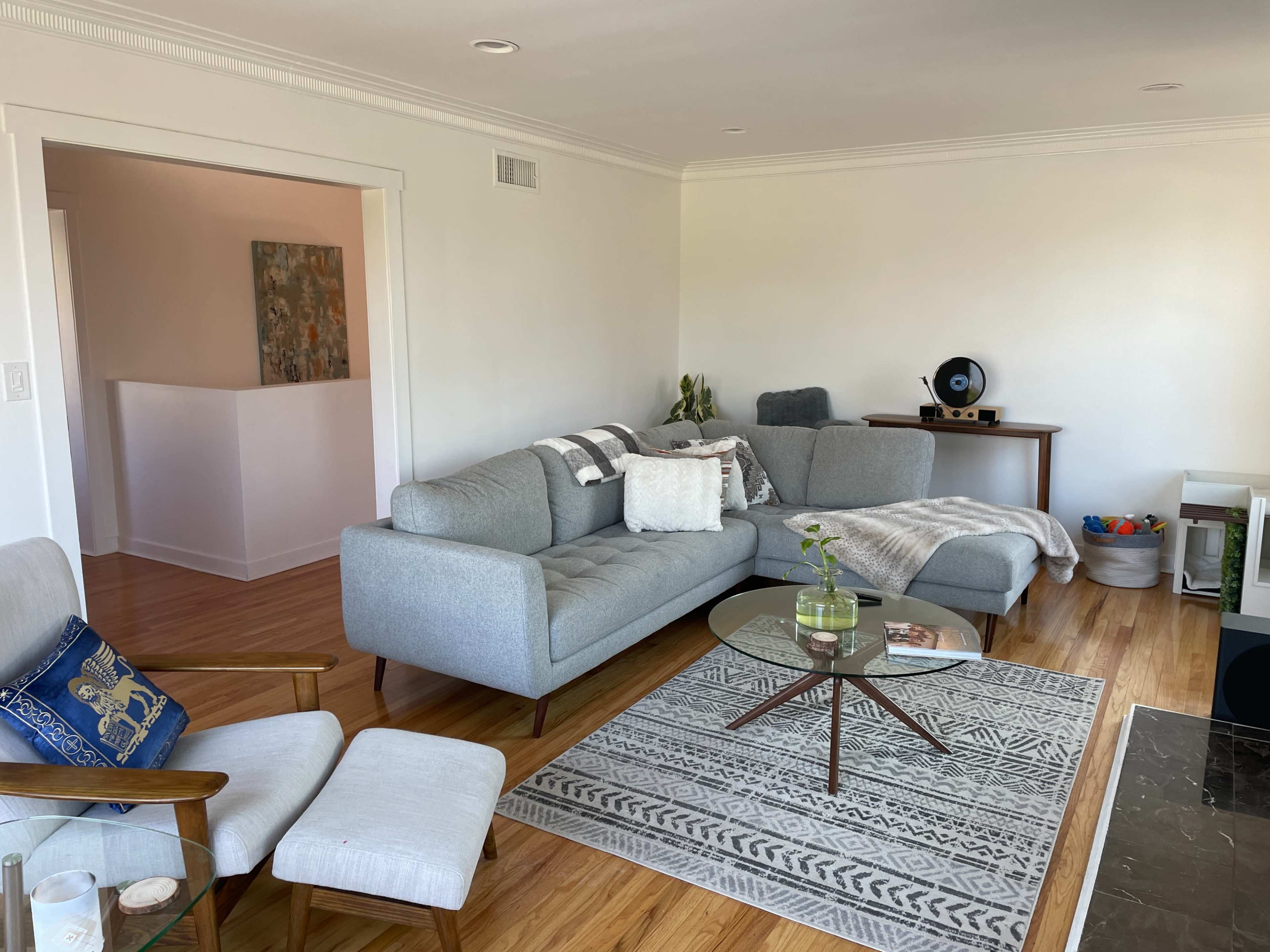 The image shows a modern living room with a light gray sectional sofa, a round glass coffee table, and a patterned area rug, complemented by wooden flooring and minimal decor.
