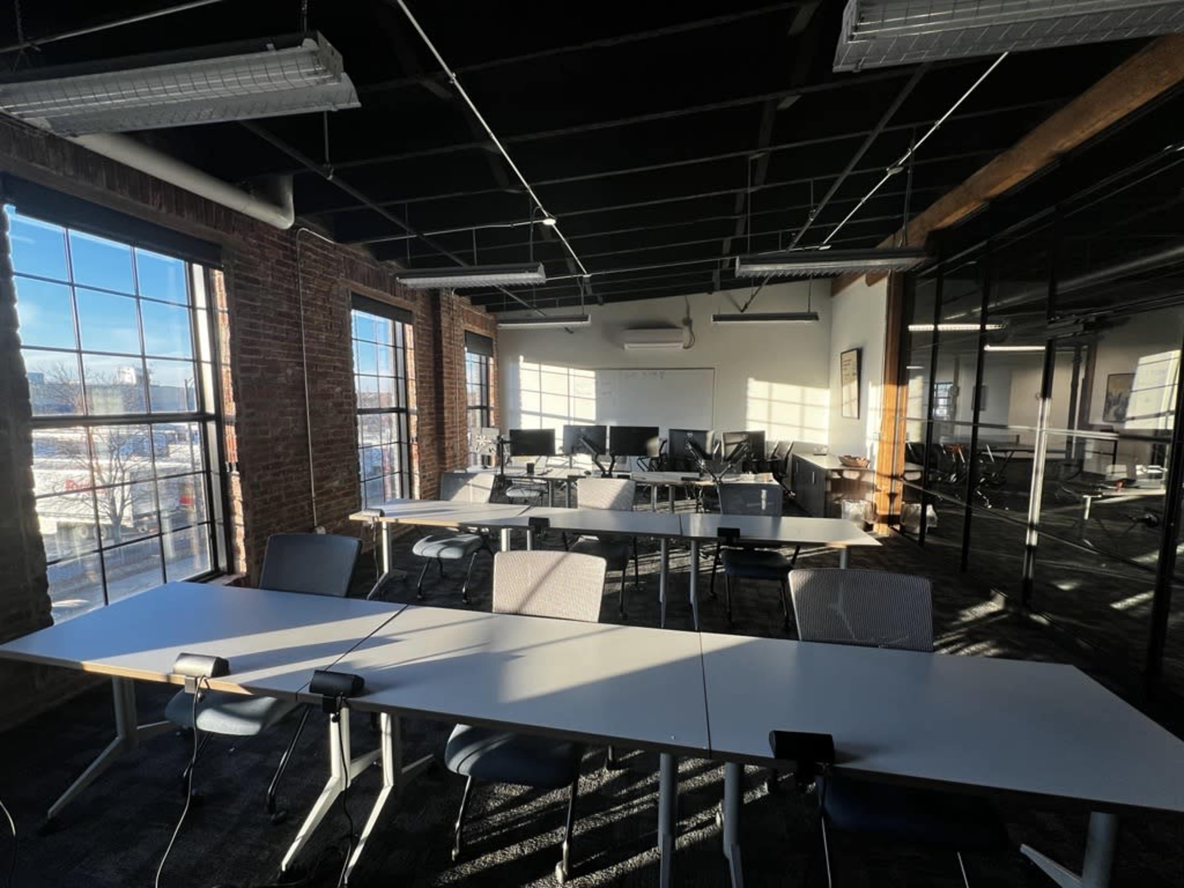A bright, modern classroom with rows of tables and chairs arranged in a spacious area featuring large windows and exposed brick walls.
