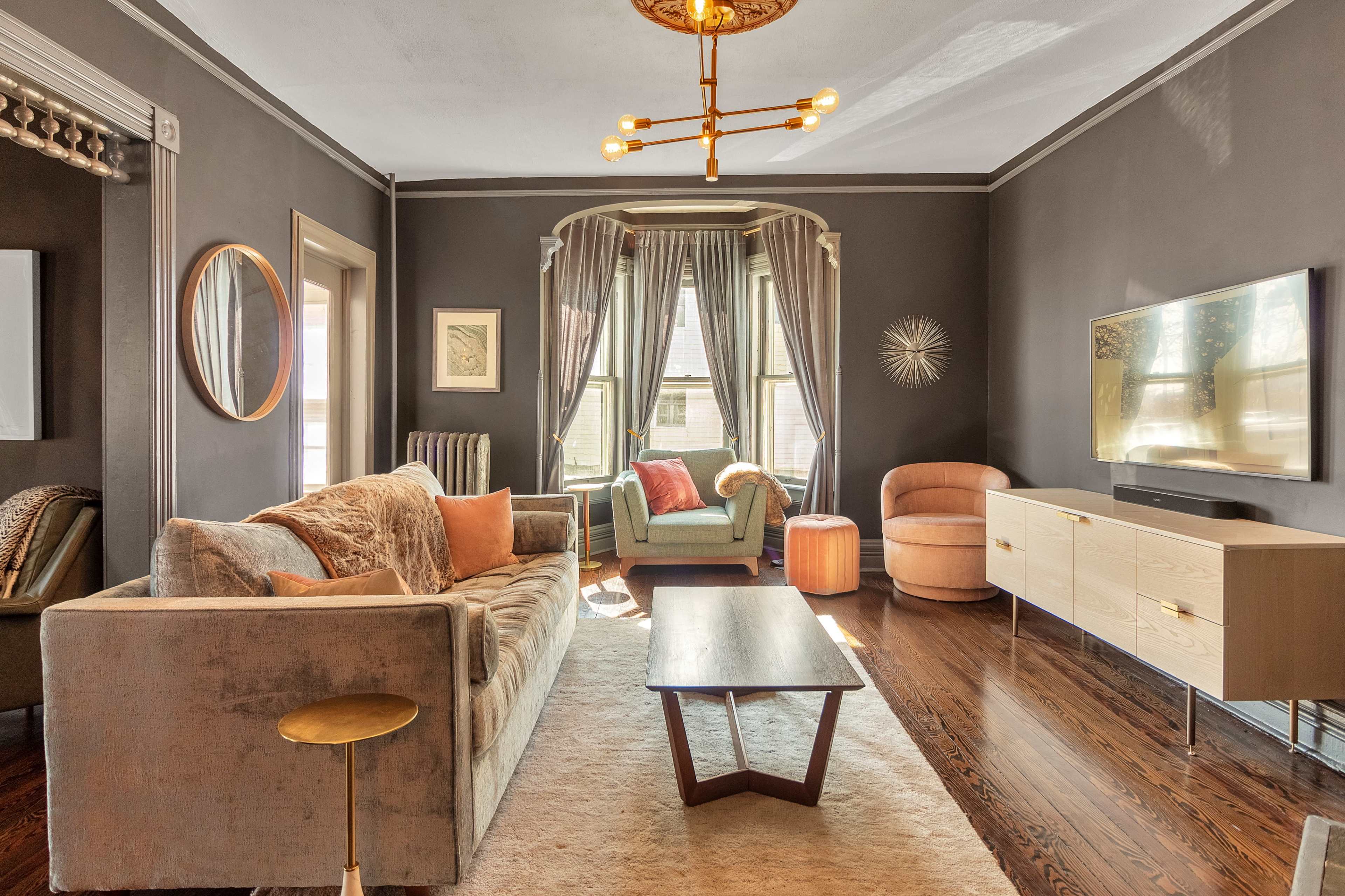 A living room features a neutral sofa and armchairs, a wooden coffee table, and a television mounted on the wall, with large windows draped in curtains.