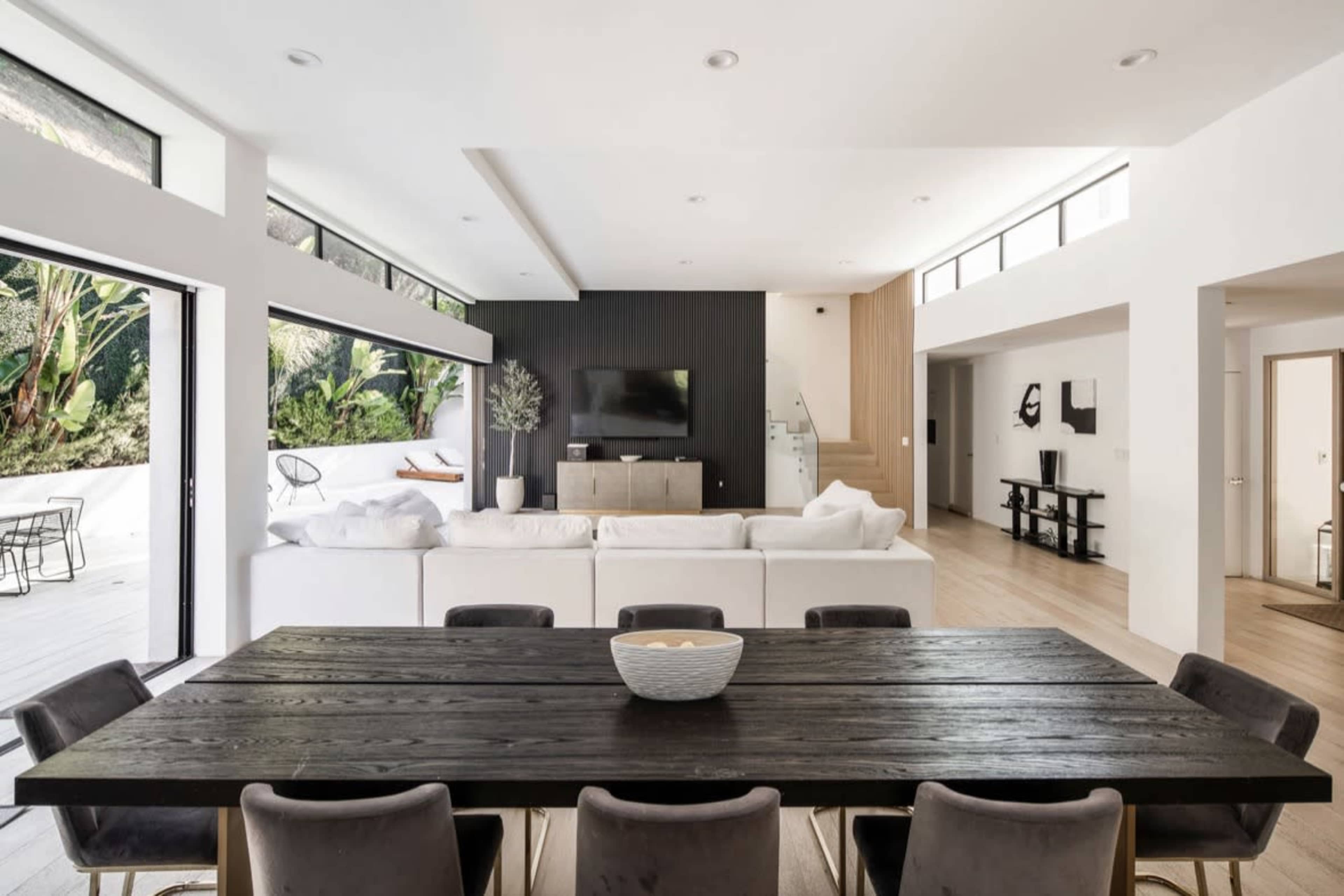 A modern, open-concept living and dining area features a large wooden dining table, white sofas, and large windows that invite natural light.