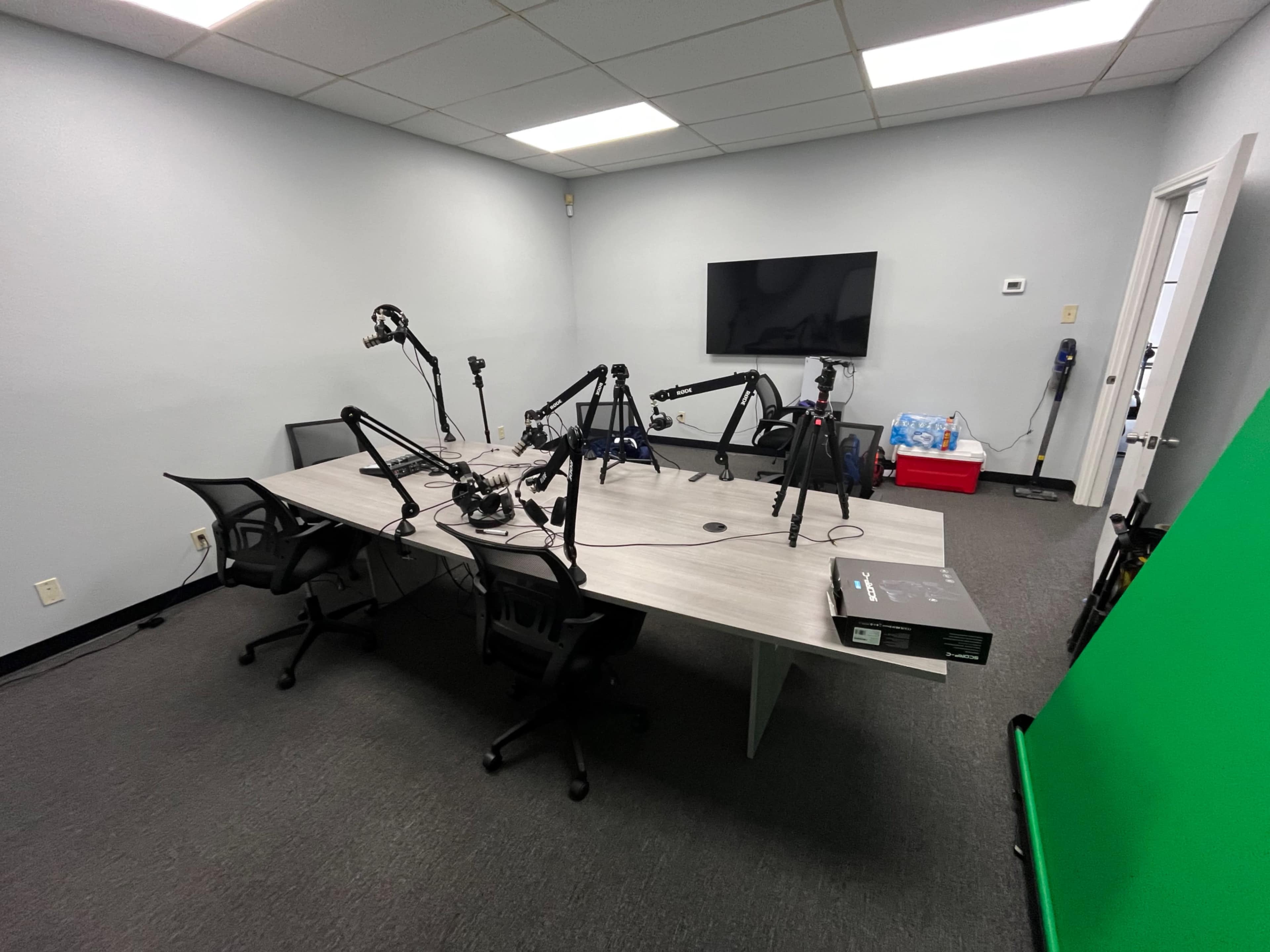 The image shows a modern recording studio with a large table, multiple microphone setups, and a green screen in one corner.