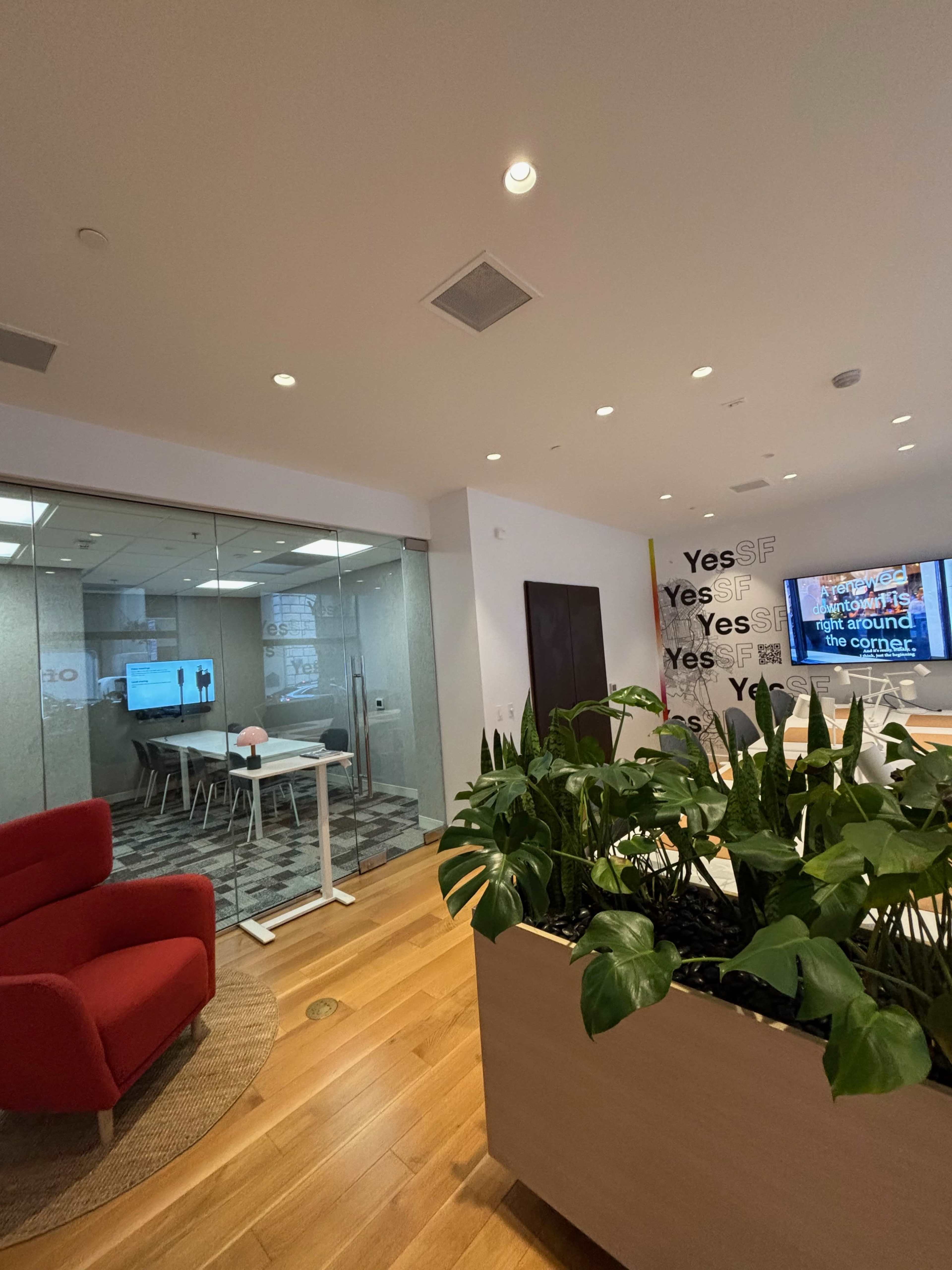 The image shows a modern office space with glass-walled conference rooms, a red chair, and indoor plants in a wooden planter.