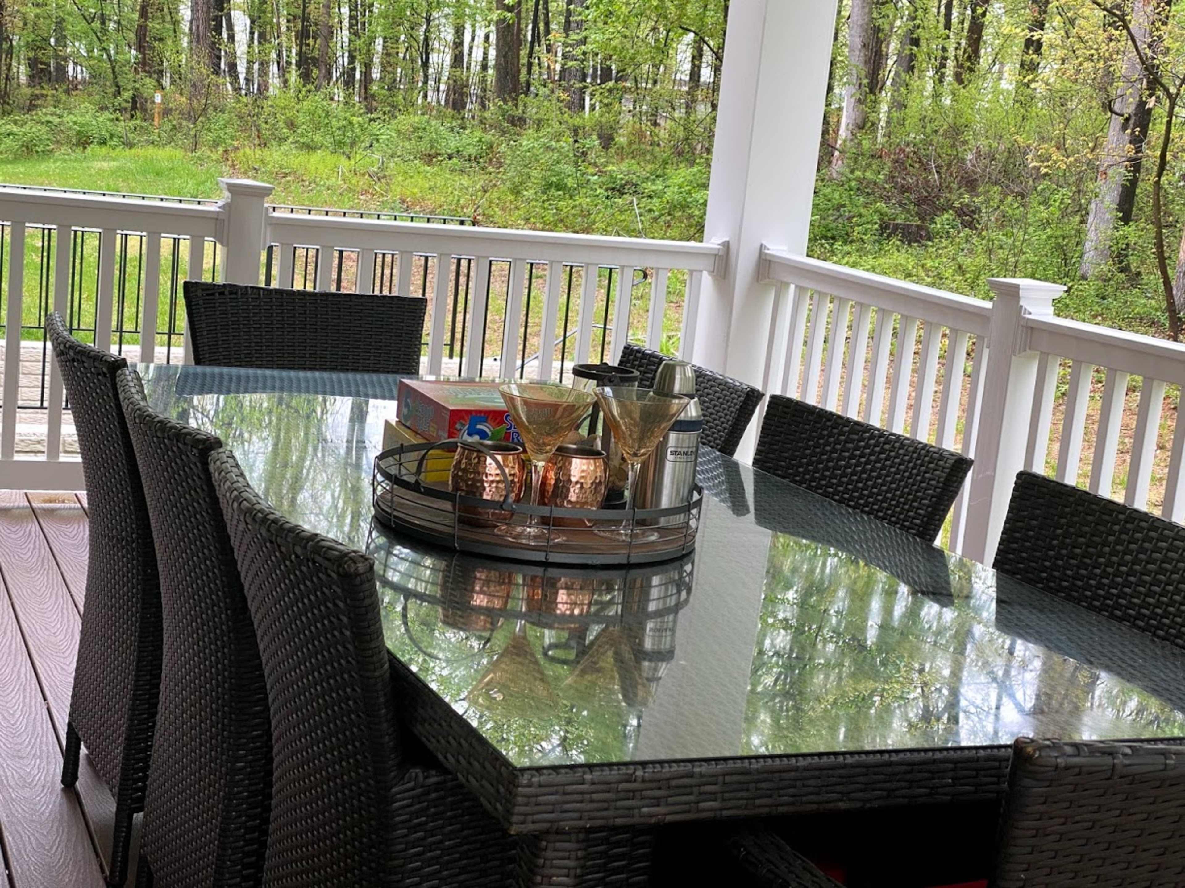 A dining table with a glass top and wicker chairs is set up on a porch overlooking a green forest.
