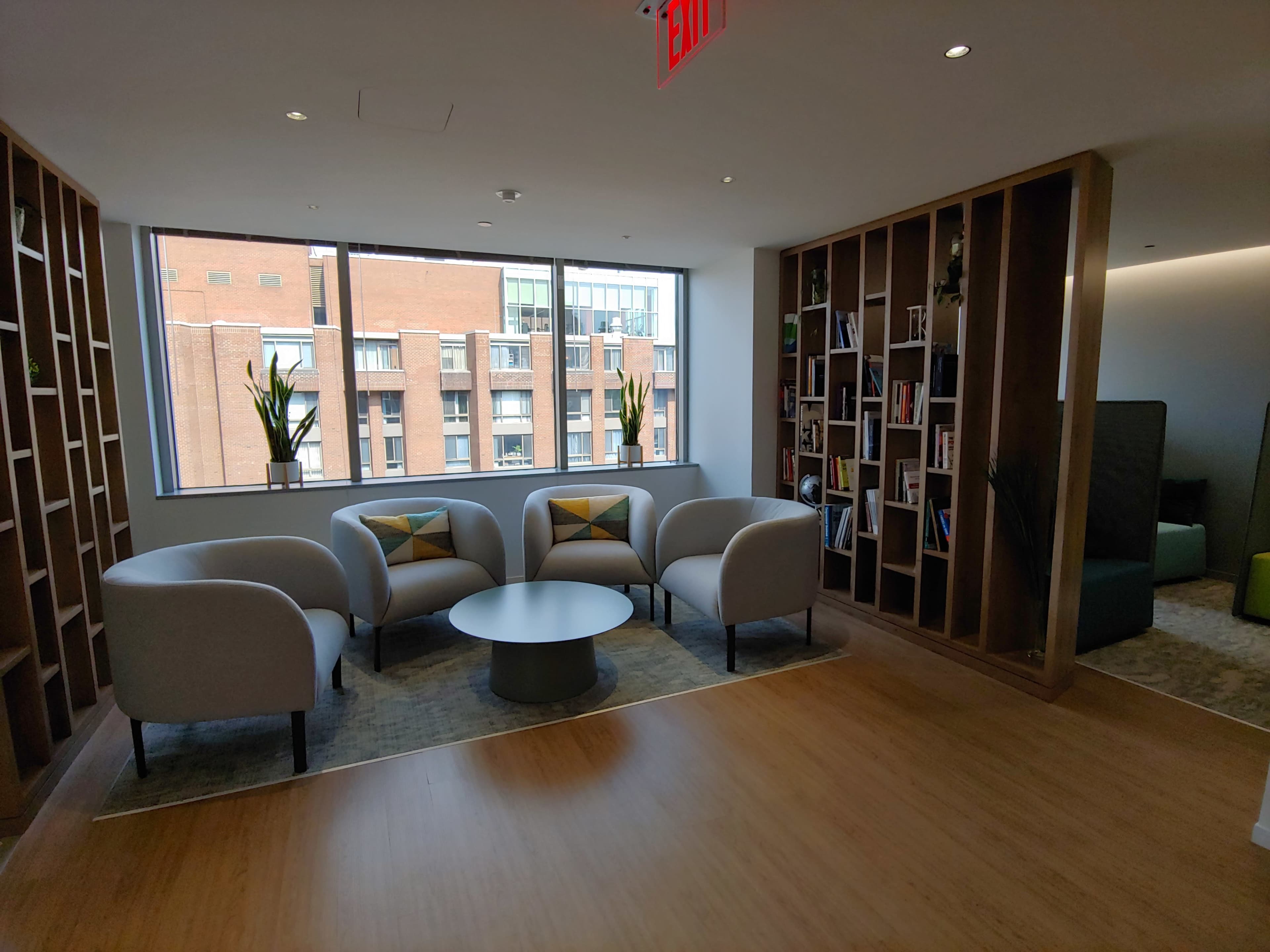 A seating area with four armchairs and a round table is arranged beside a window displaying a brick building exterior.