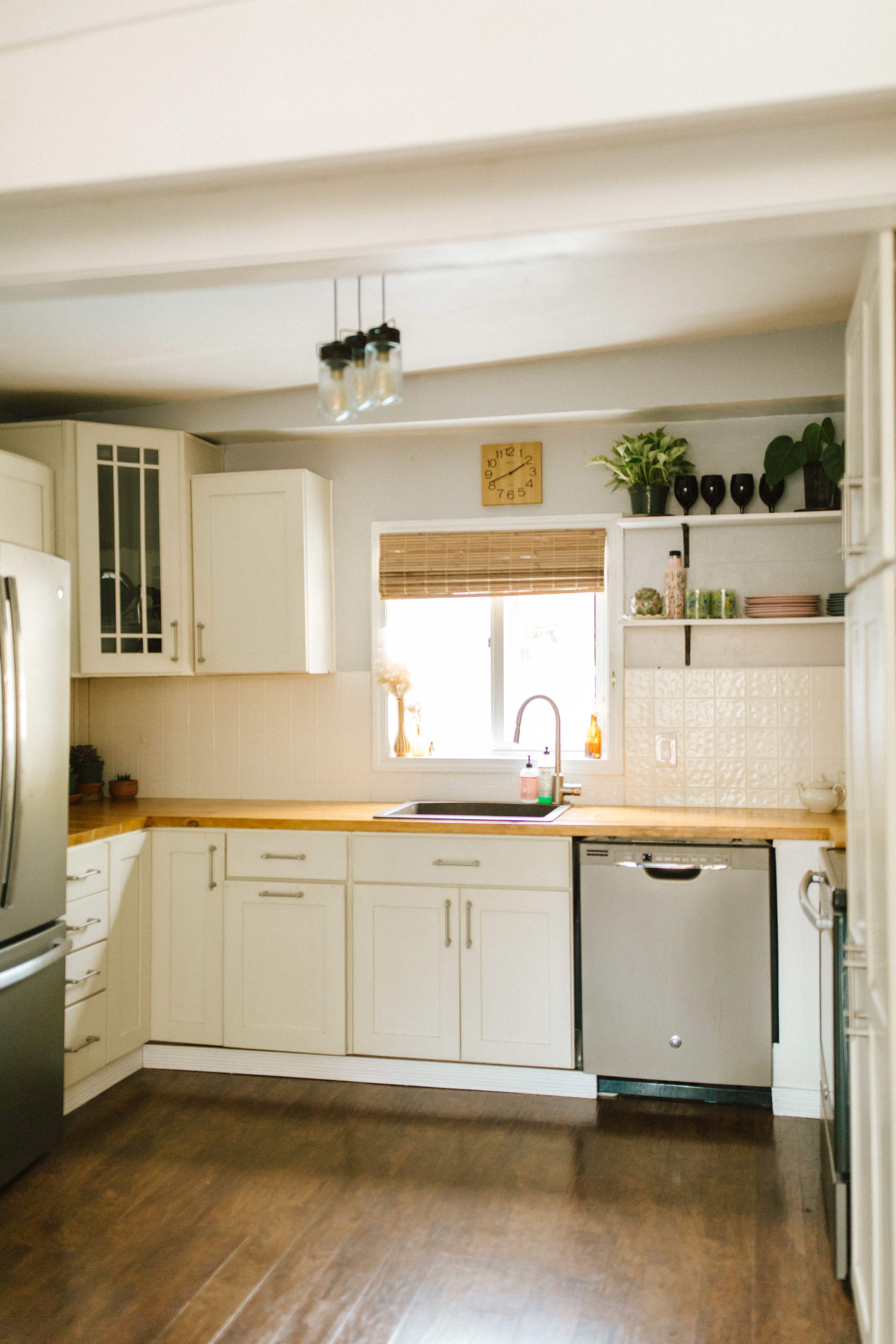 A bright kitchen features cream-colored cabinets, a stainless steel refrigerator, and a sink below a window with a bamboo shade.