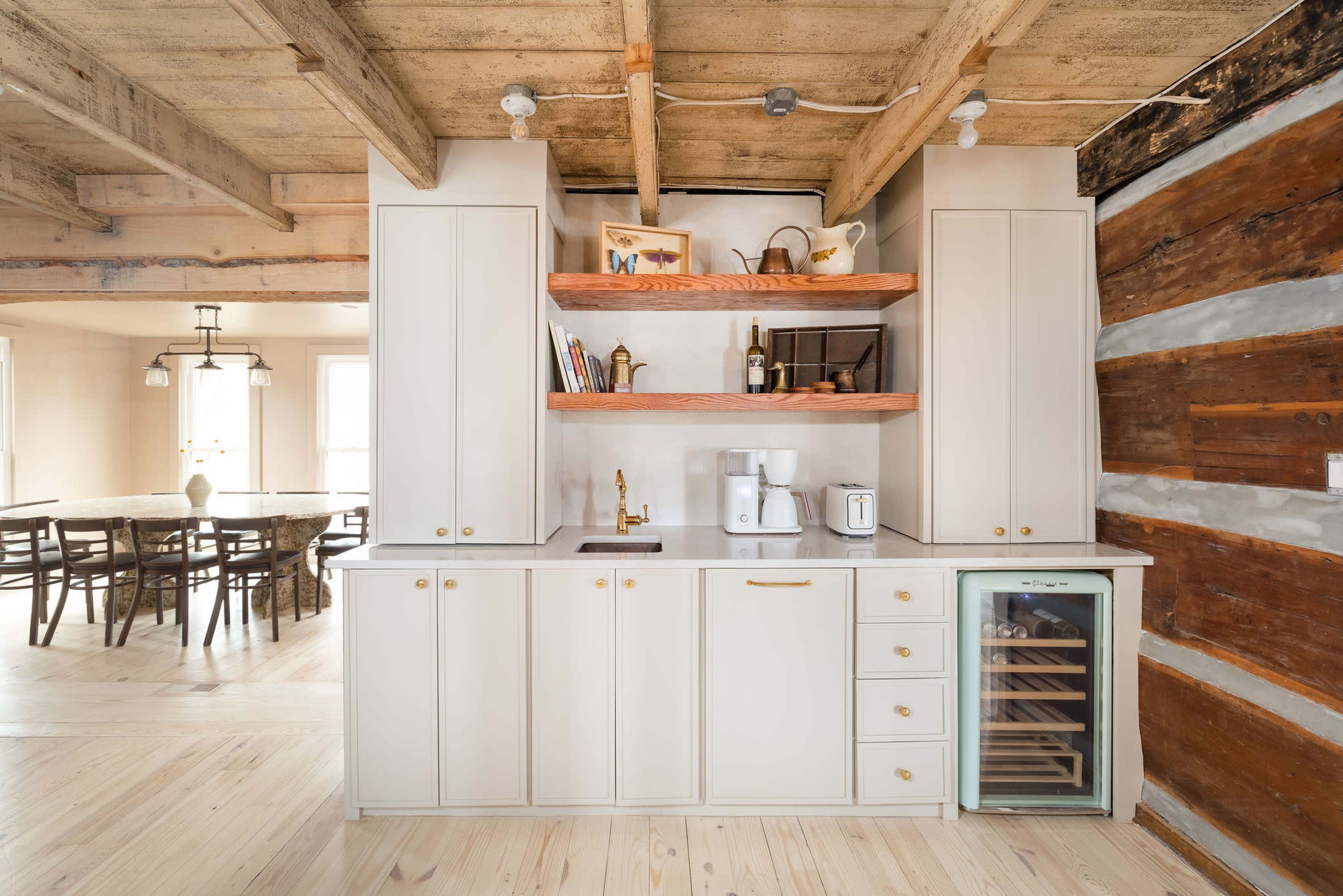 A modern kitchen with light-colored cabinetry, open shelving, and a wooden accent wall, alongside a dining area in the background.