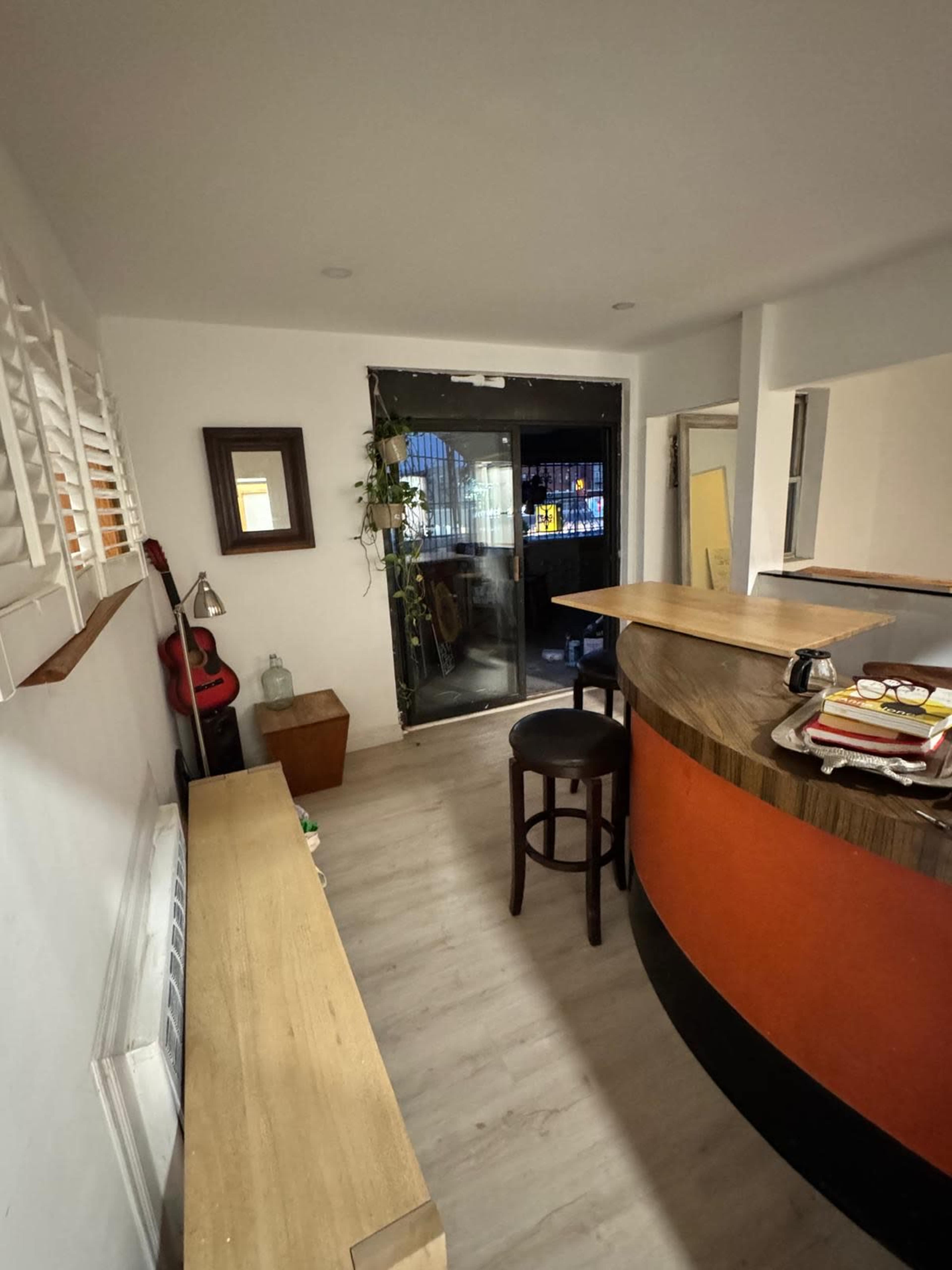 The image shows a modern interior space with a wooden counter, a stool, and a glass door leading to an outdoor area.