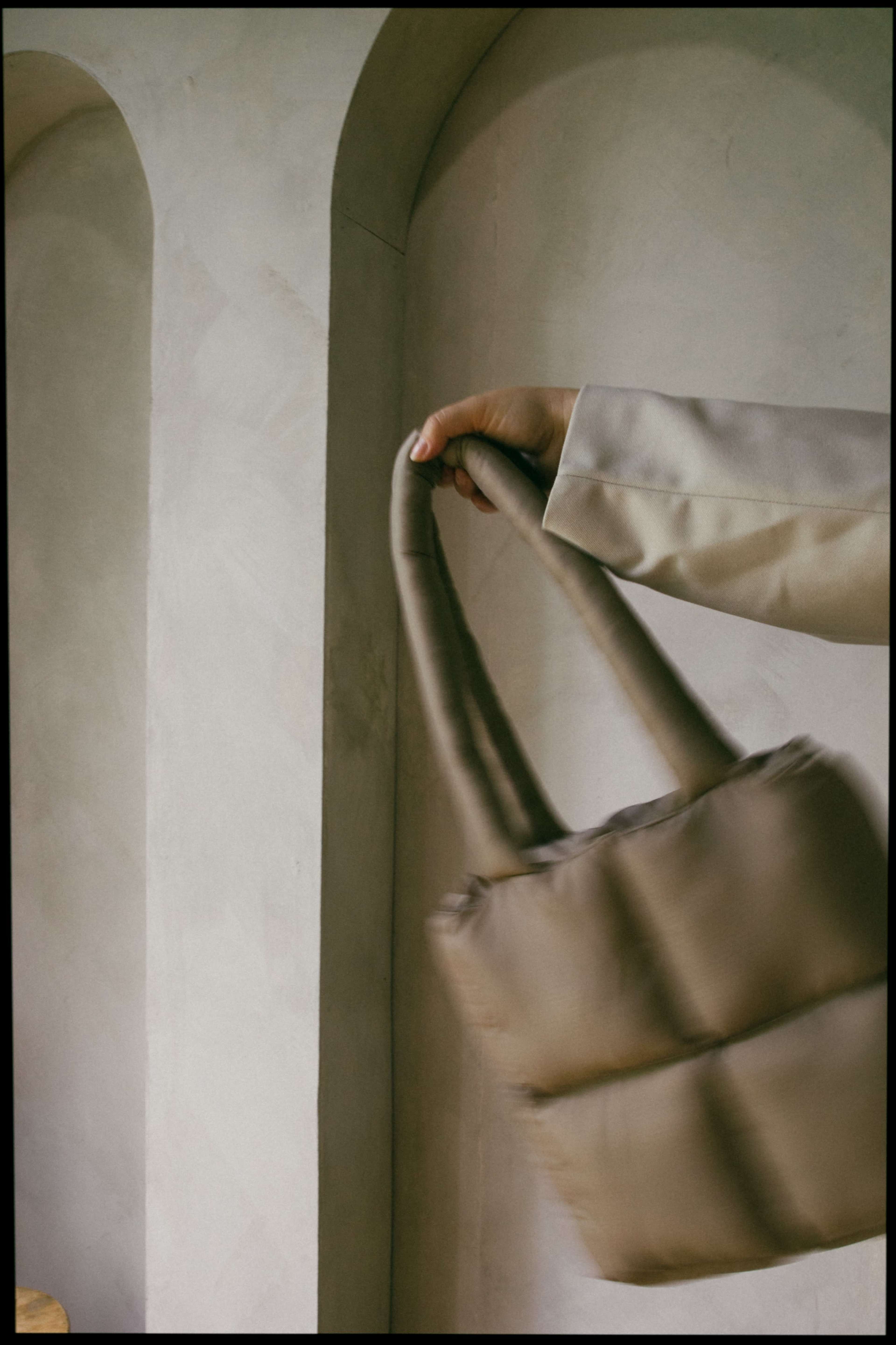 A hand holds a beige, padded tote bag near a textured wall with arched architectural details.