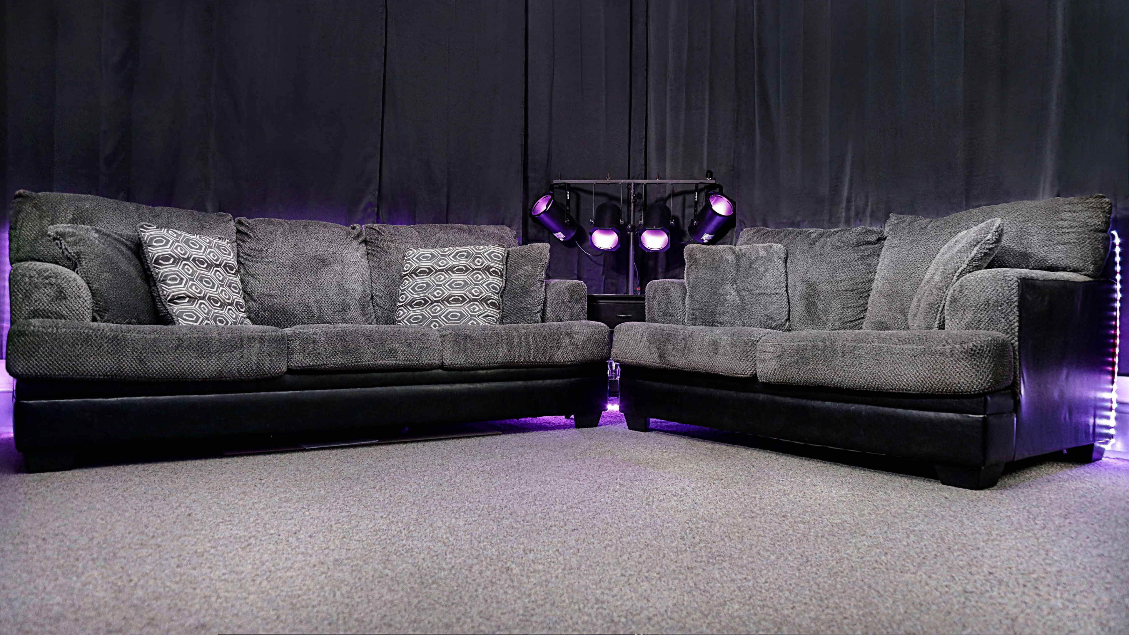 Two grey fabric sofas positioned at an angle, with decorative cushions on one and purple stage lights in the background.