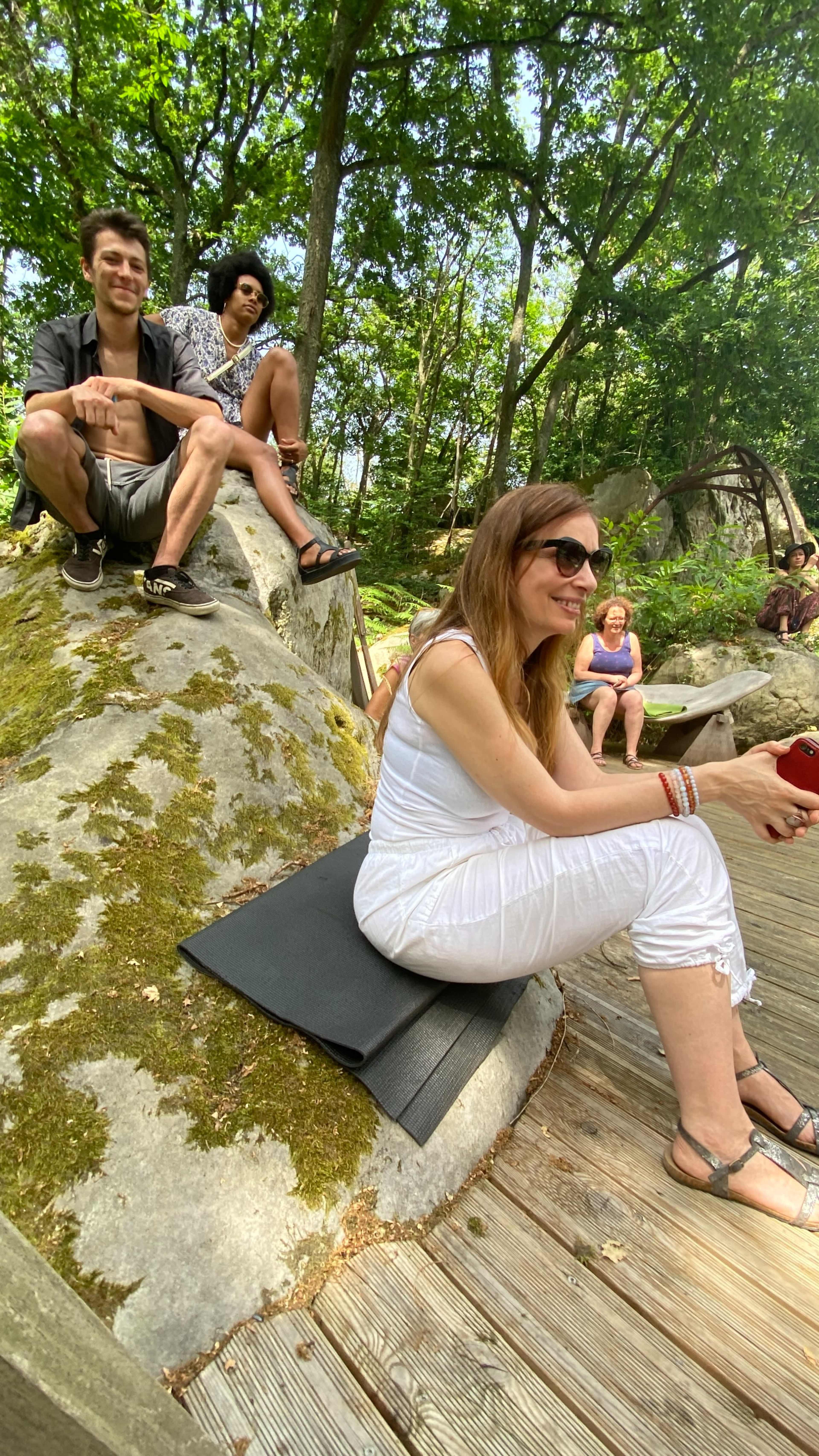 Three people relax on a moss-covered rock in a wooded area, while others sit nearby on a wooden platform.