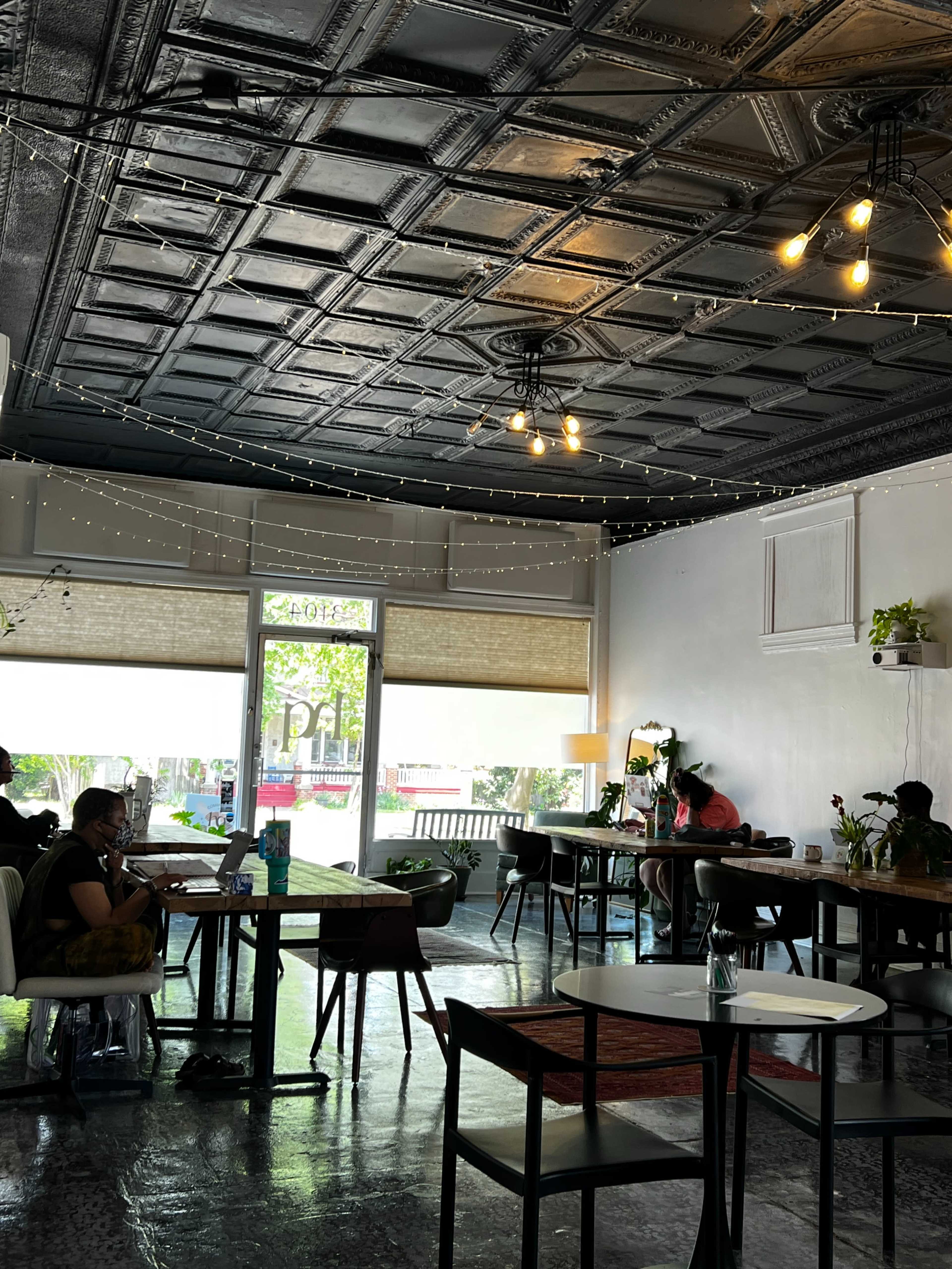 The image shows a modern café interior with tables and chairs, featuring a decorative ceiling and string lights, with a few patrons engaged in work and conversation.