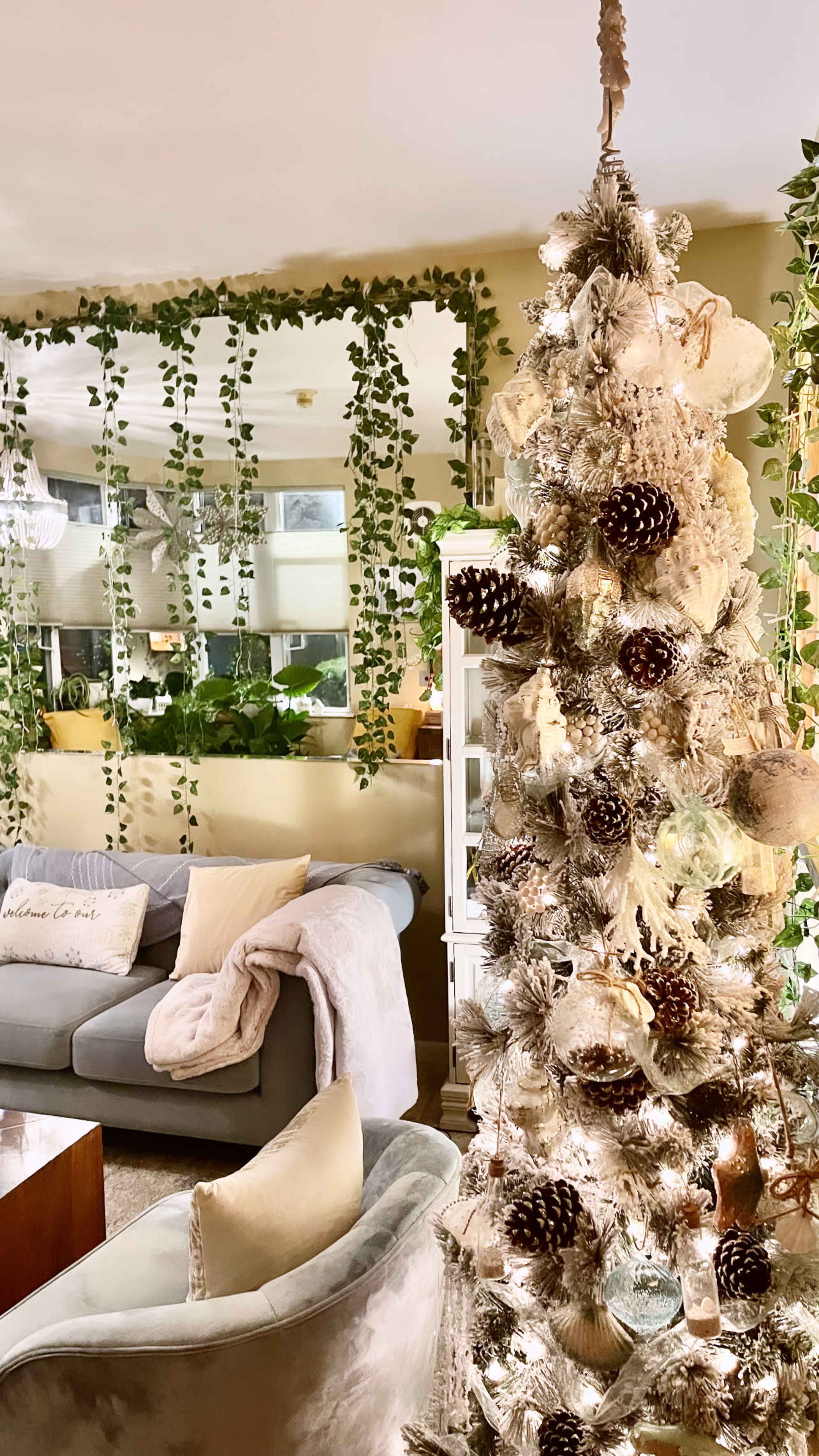 A decorated Christmas tree adorned with pinecones and lights stands in a living room surrounded by green vines and comfortable furniture.