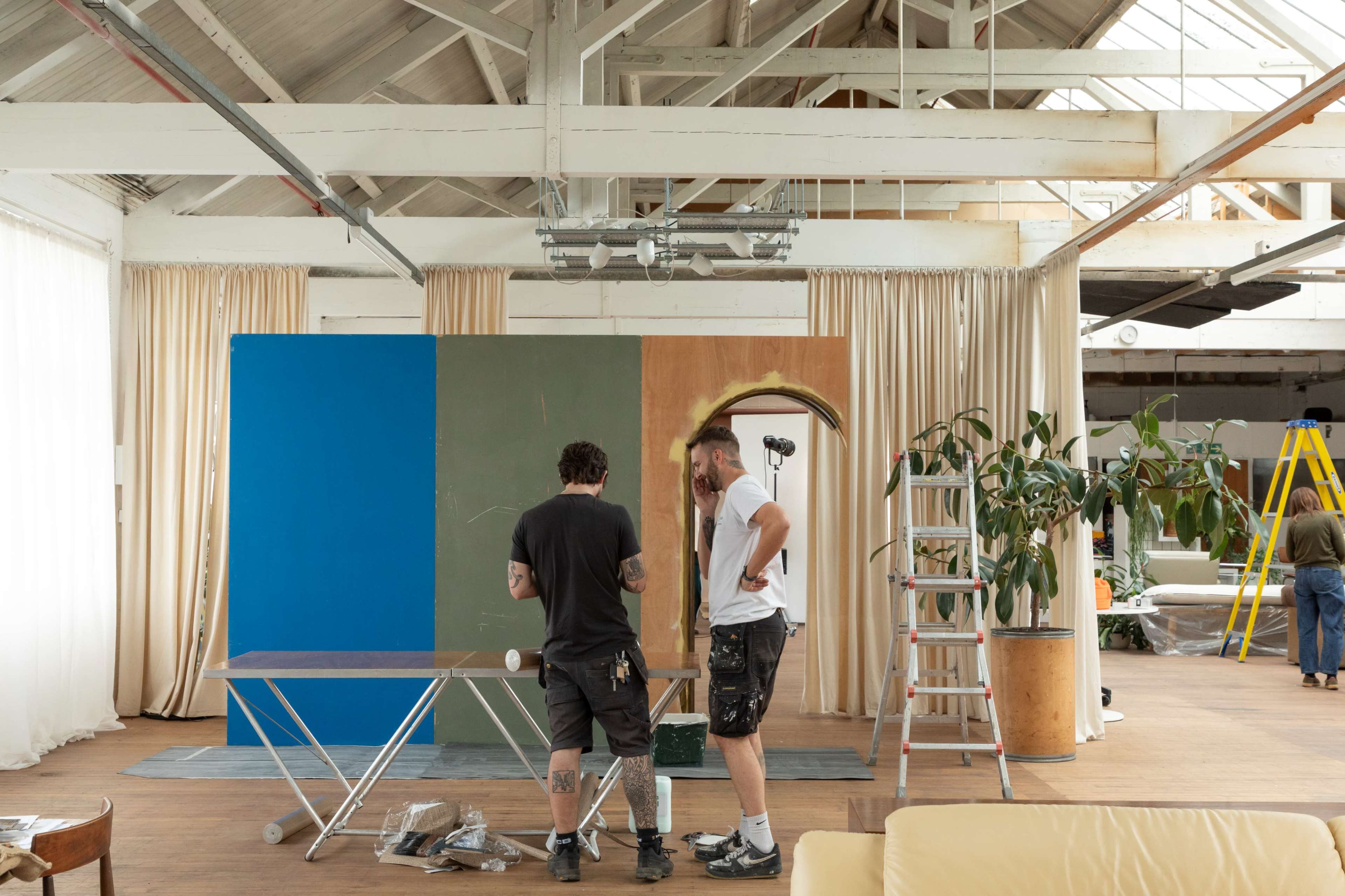 Two men are discussing in a spacious room with high ceilings, surrounded by tools and materials for painting a colorful backdrop.