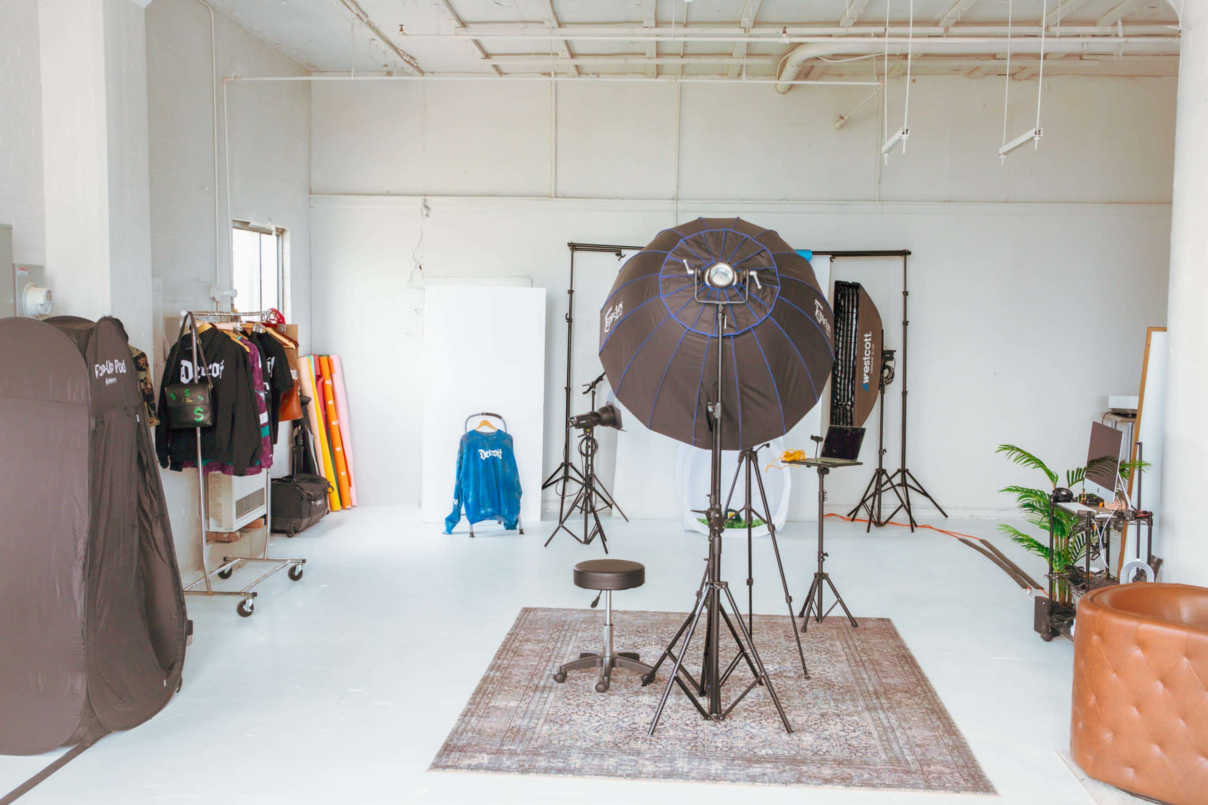 A photography studio features a lighting setup, a stool, various backdrops, and clothing racks along with a decorative rug and a plant.