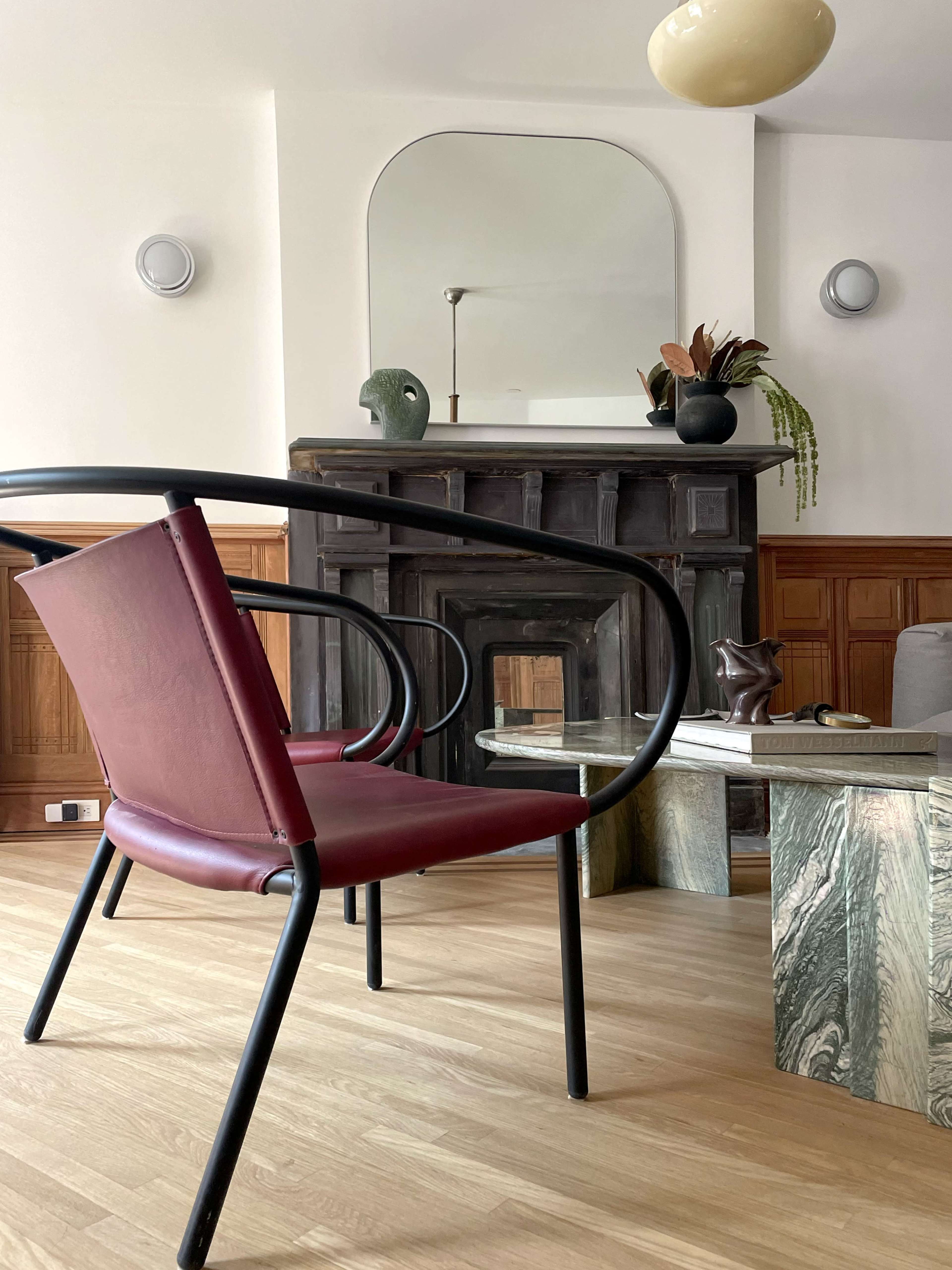 A modern chair is positioned in front of a dark wood fireplace, with a mirrored wall and decorative elements in a spacious room.