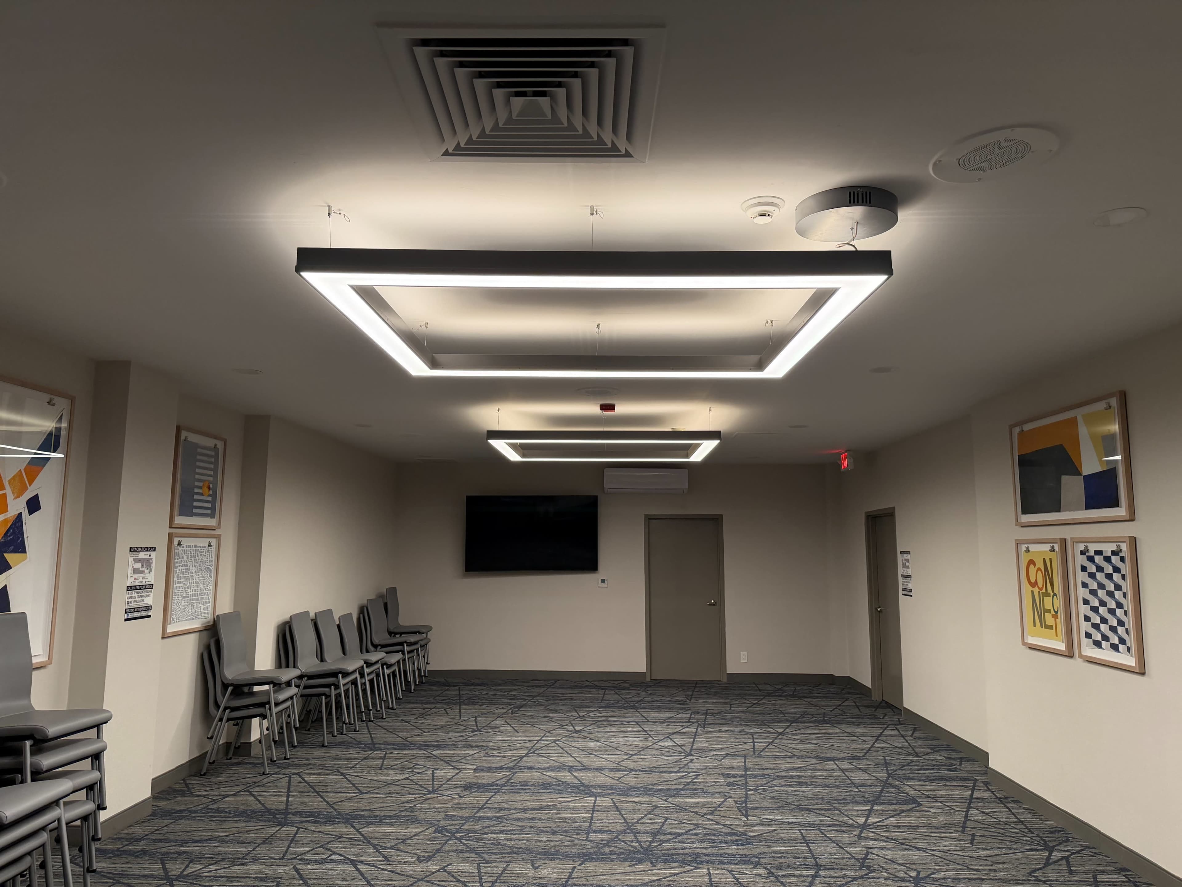 The image shows a modern indoor hallway featuring geometric ceiling lights, a television on the wall, and neatly stacked chairs along the side.