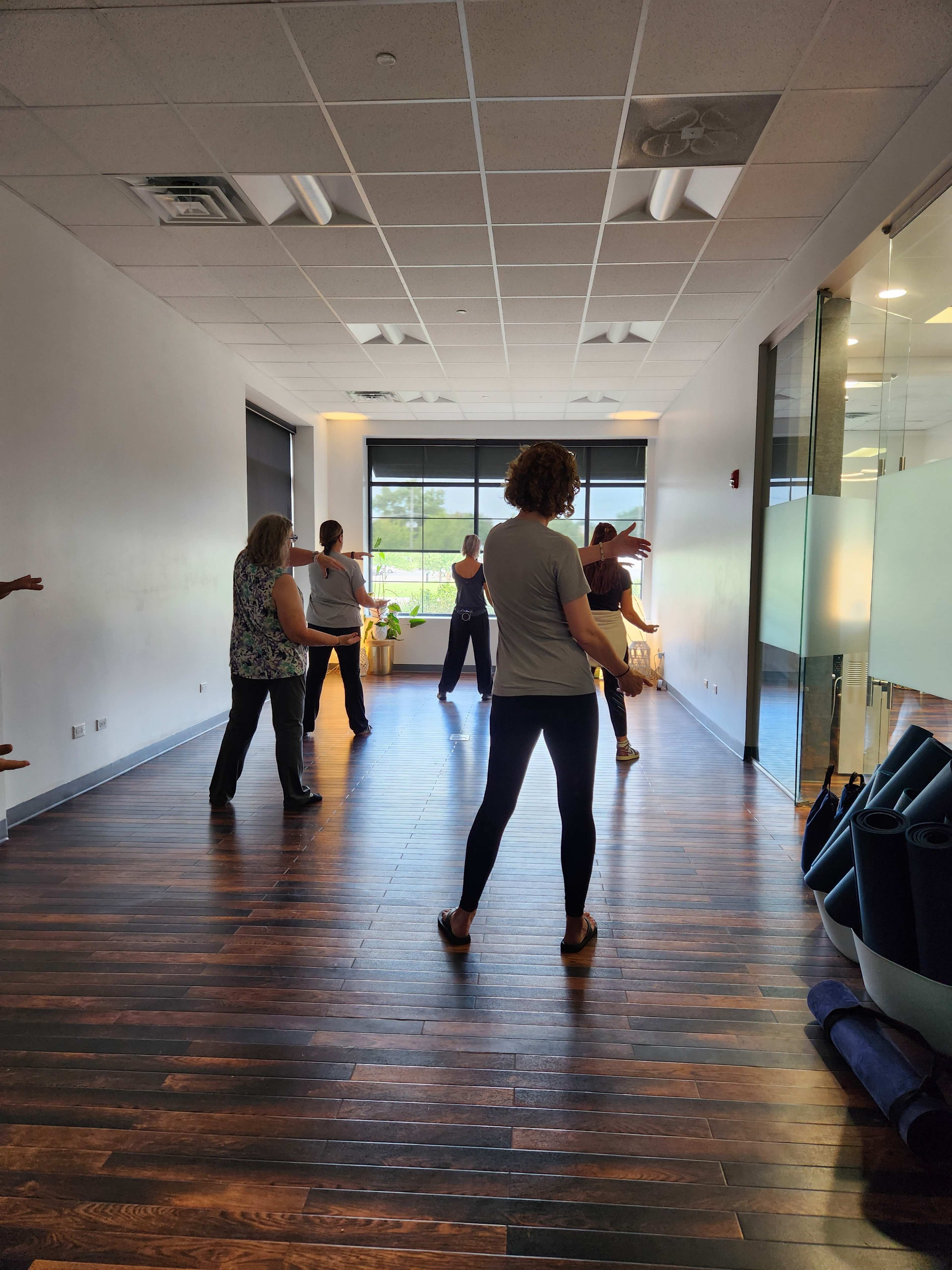 A group of people is practicing yoga or tai chi in a spacious, well-lit studio with wooden flooring and large windows.